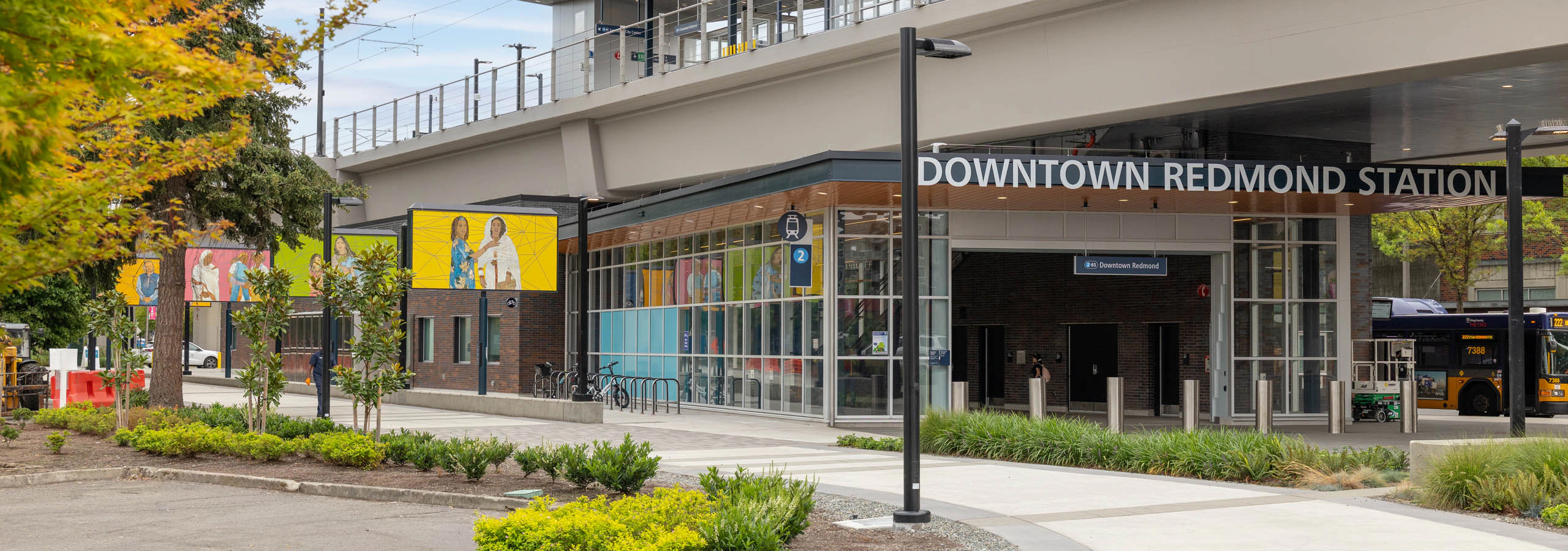 Downtown Redmond Station entrance with colorful mural artwork, landscaping, and the elevated light rail platform above, located near AMLI Redmond Way.