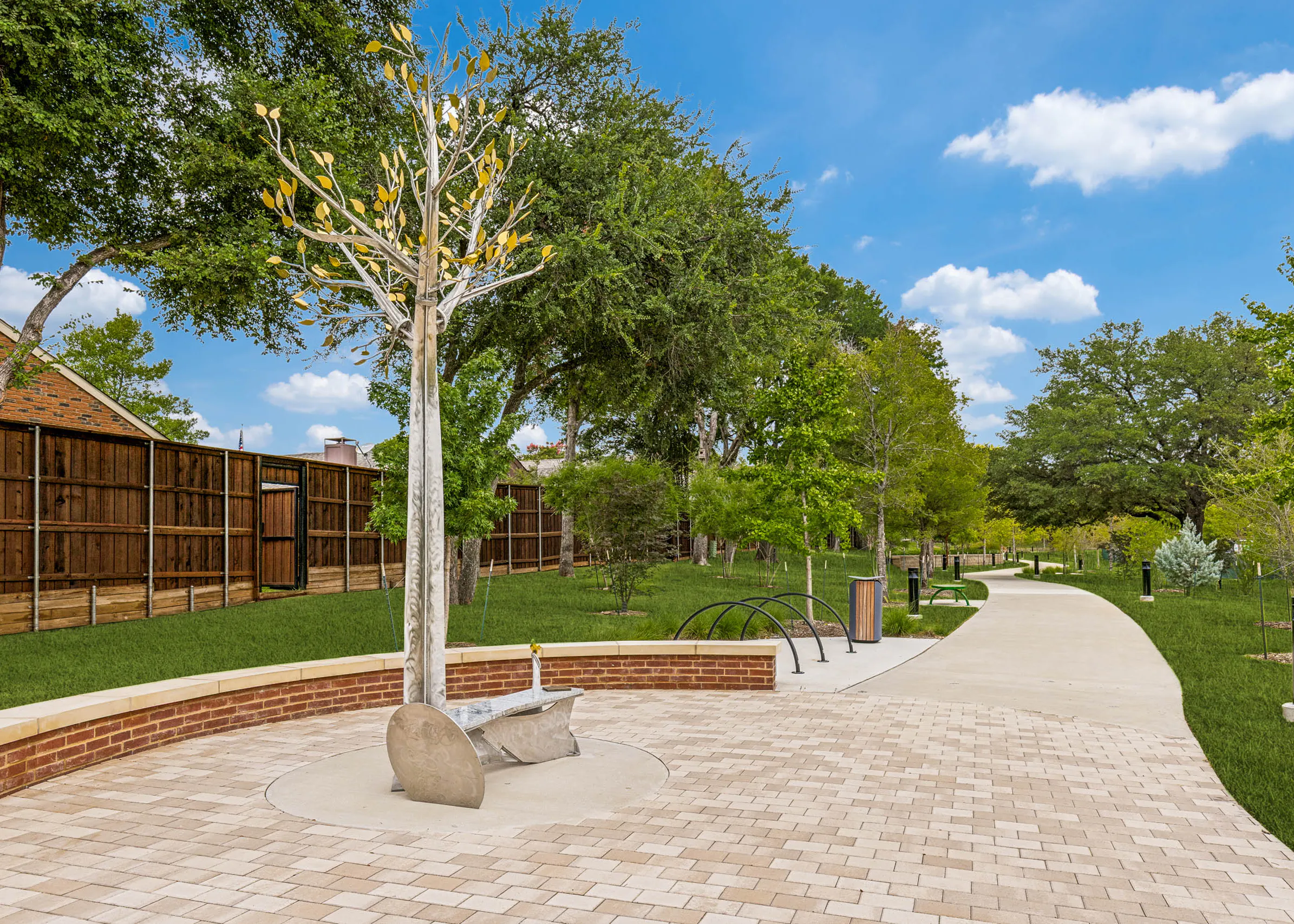 Park-like community trail featuring a decorative tree sculpture, paved seating area, landscaped greenery, pathway lighting, and open green space at AMLI Treehouse.