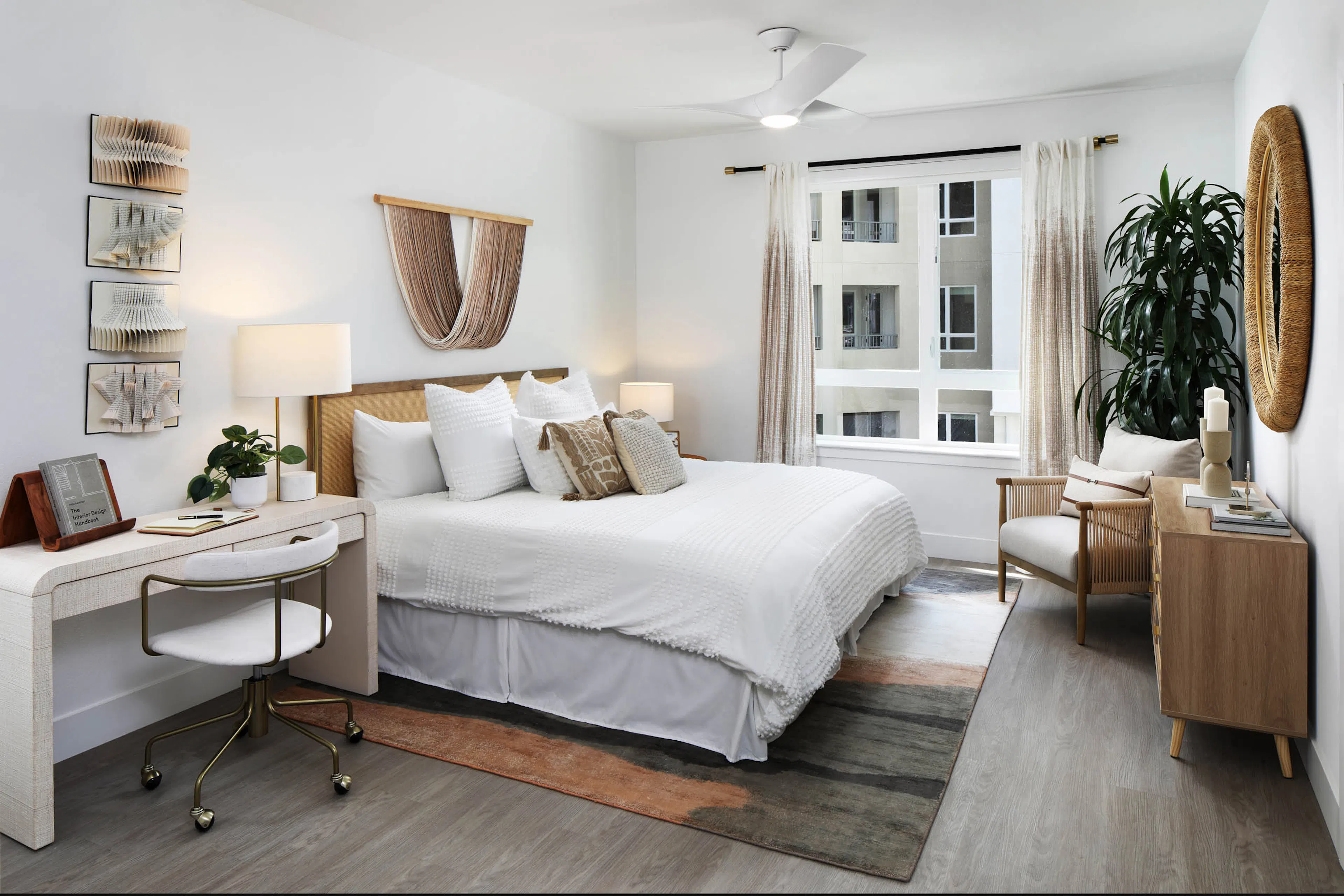 Bright bedroom with white bedding, woven headboard, desk, chair, wall art, large window with curtains, ceiling fan, plant, mirror, and wood console.