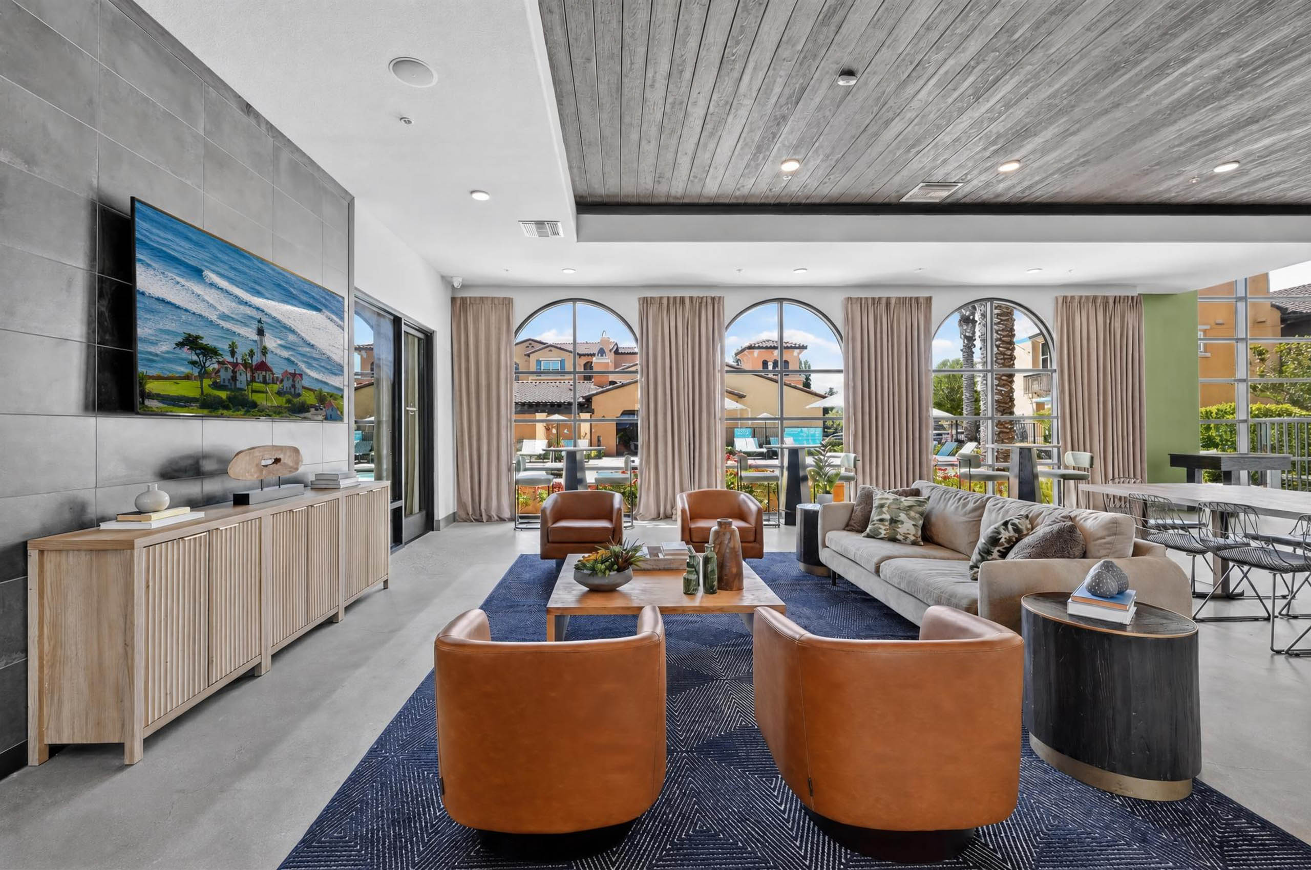 Clubhouse at AMLI Camarillo apartments with leather chairs, sofa, coffee tables, television and tall arched windows overlooking the pool.