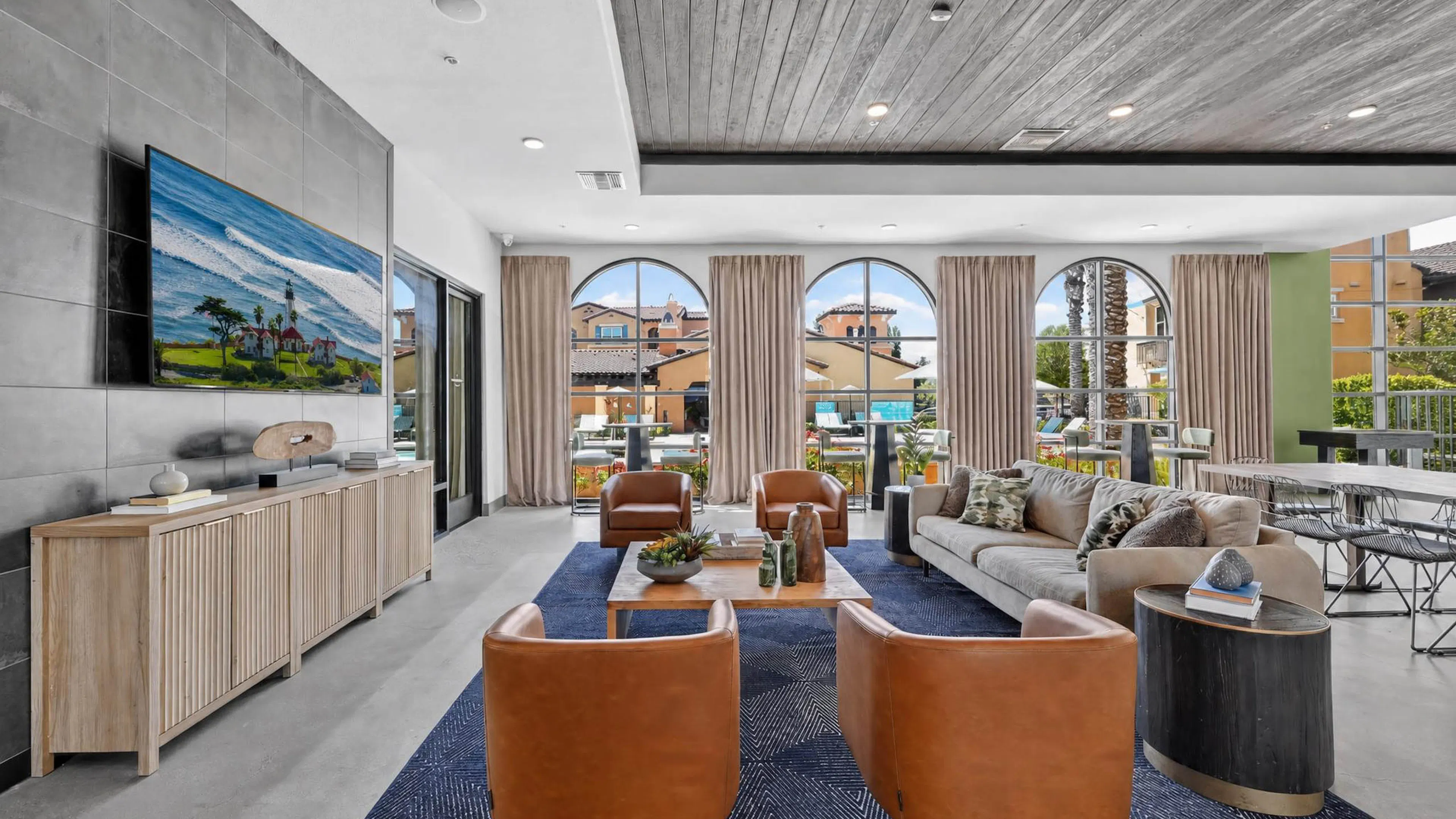 Clubhouse at AMLI Camarillo apartments with leather chairs, sofa, coffee tables, television and tall arched windows overlooking the pool.