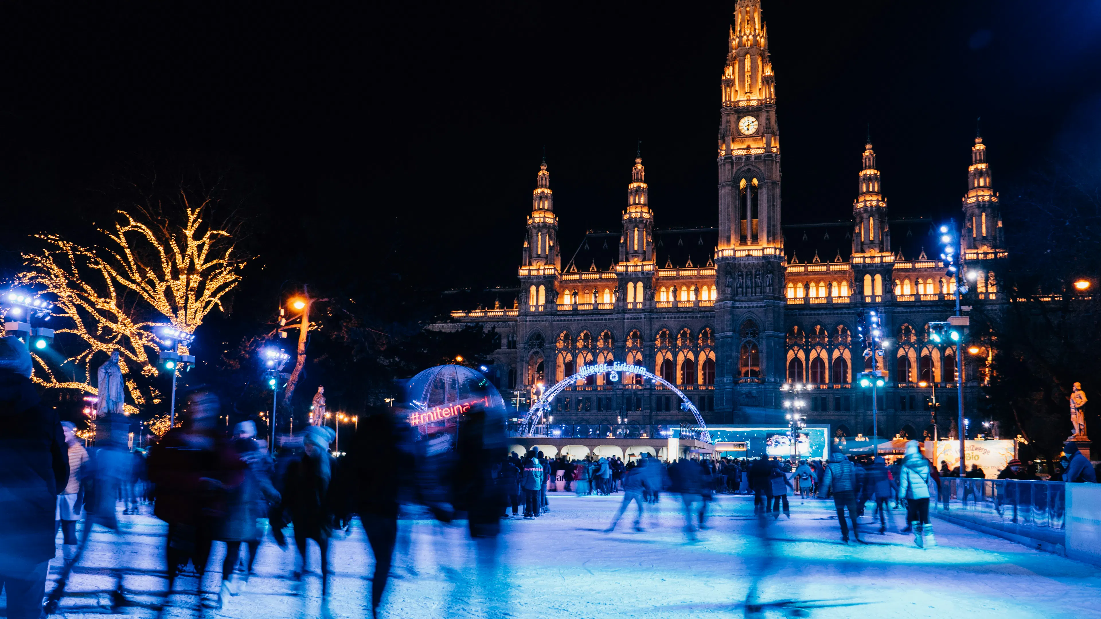 A long-exposure shot of ice skaters at an outdoor ice rink beneath a building lit with holiday lights