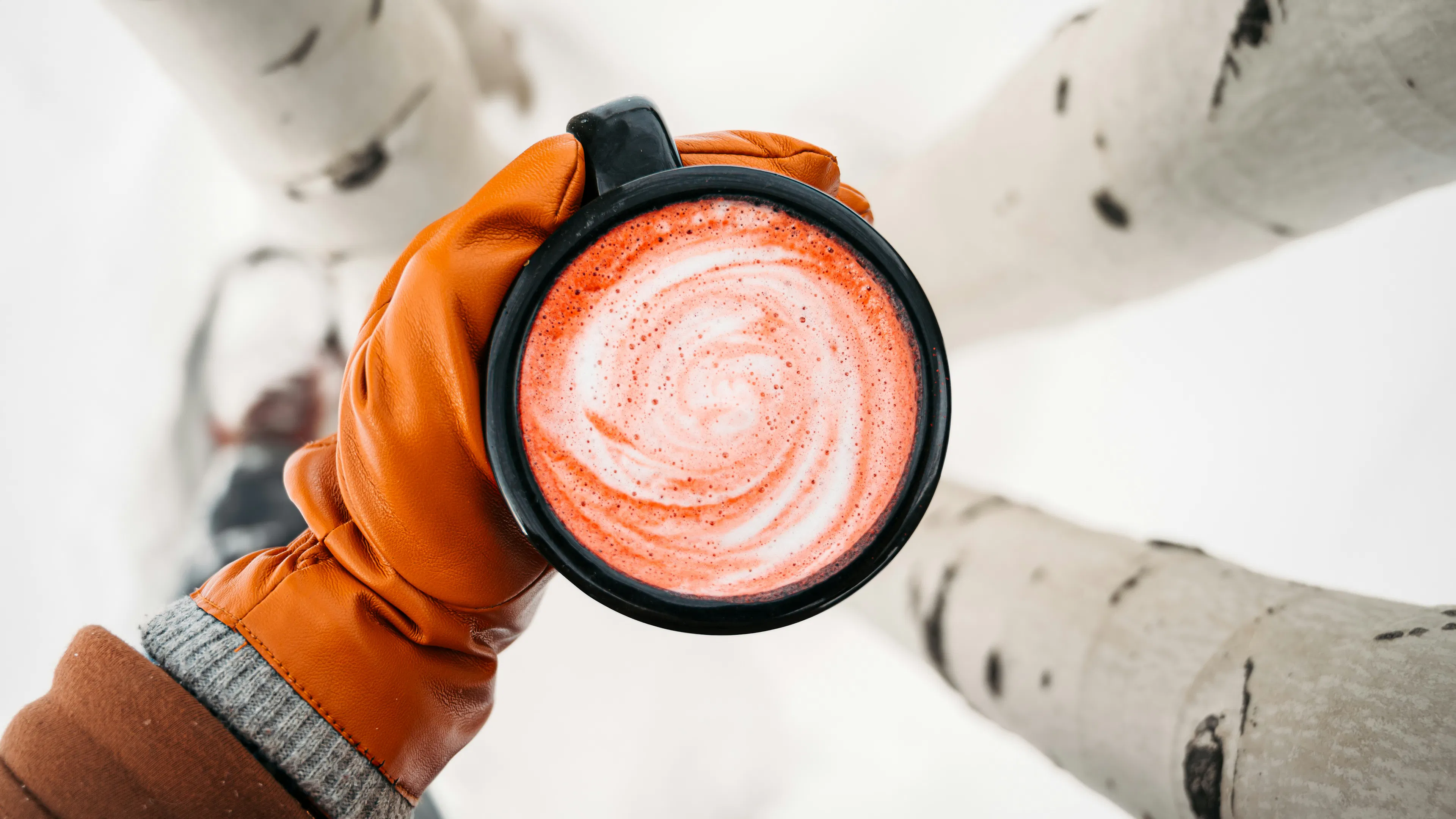 A gloved hand holding a mug of hot cocoa above snow covered ground
