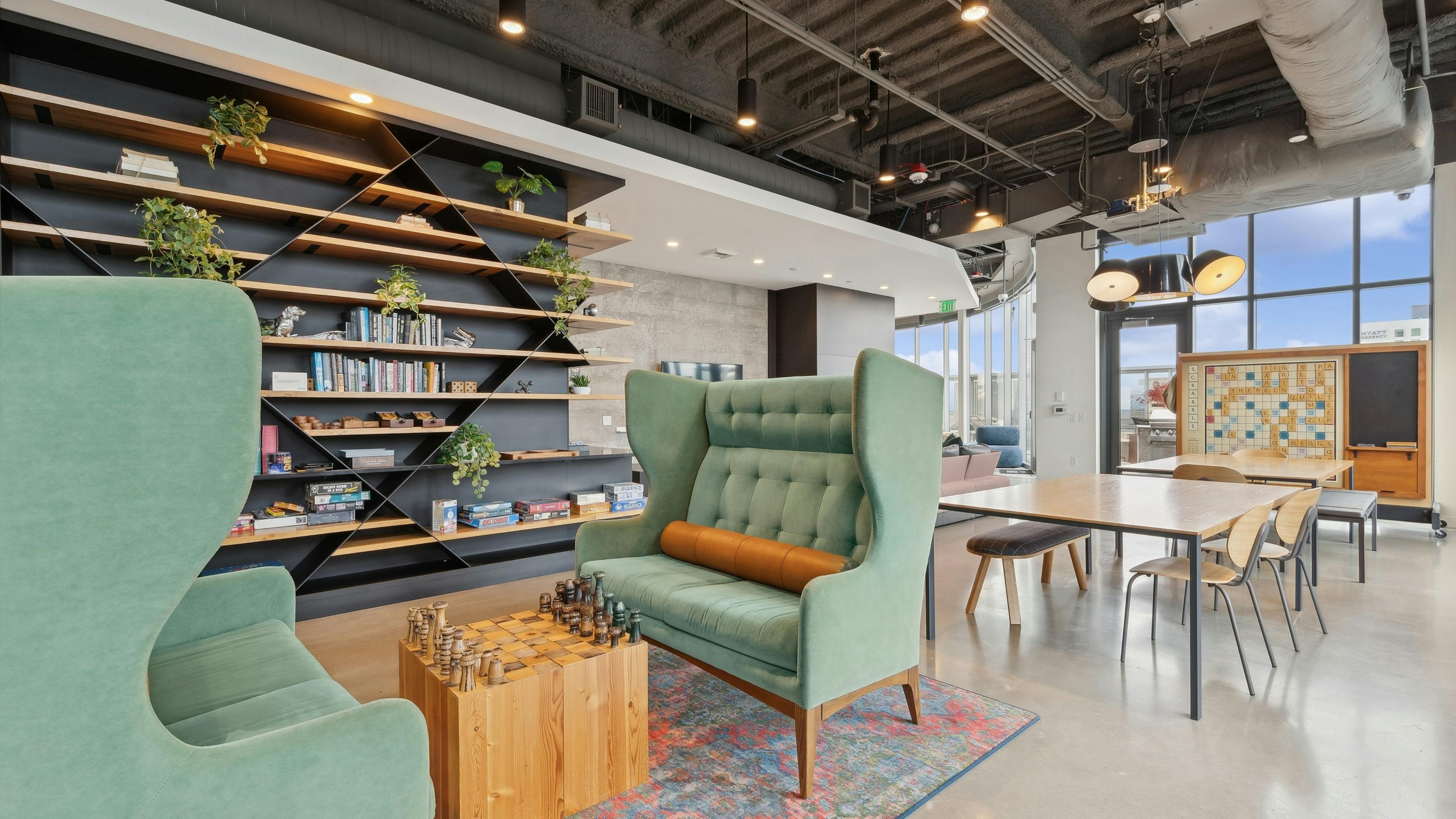 Game zone featuring green wingback chairs, a chessboard, wall shelving with books and games and a communal table at AMLI Arc