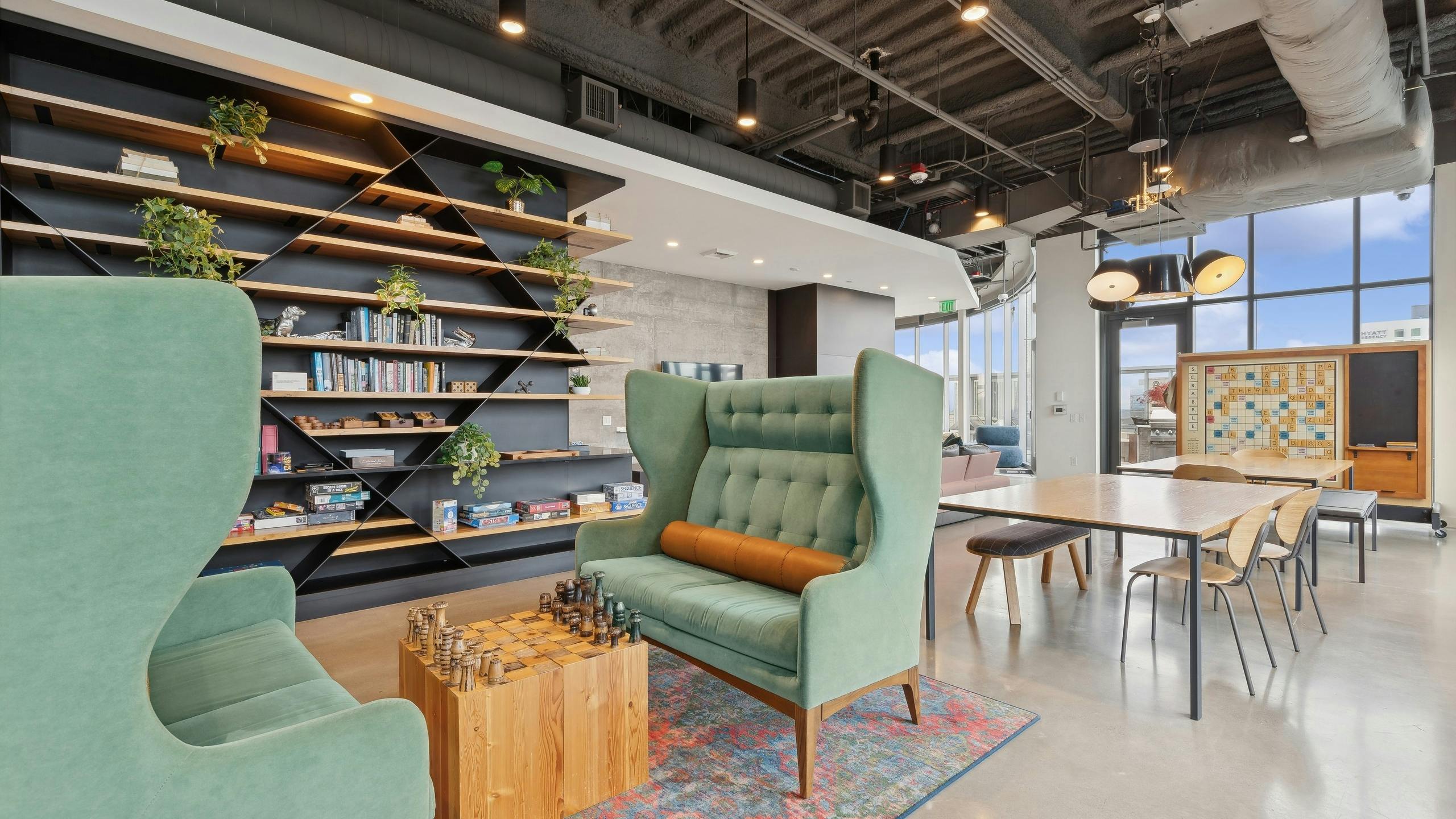 Game zone featuring green wingback chairs, a chessboard, wall shelving with books and games and a communal table at AMLI Arc