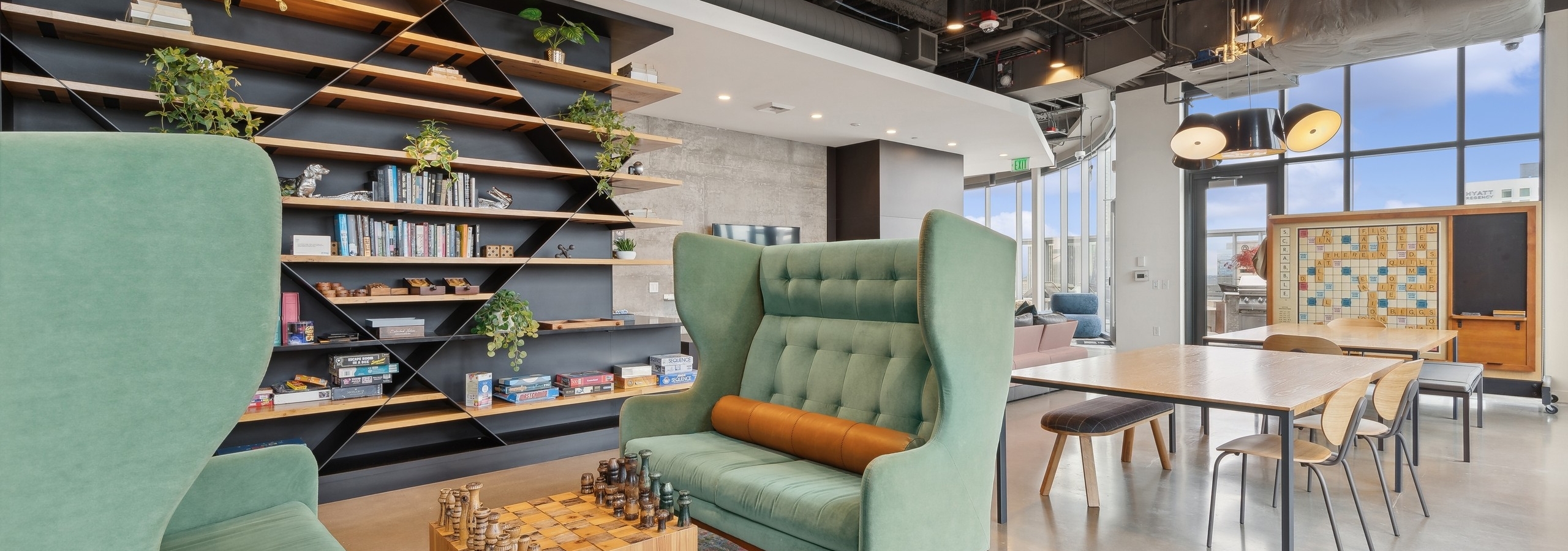 Game zone featuring green wingback chairs, a chessboard, wall shelving with books and games and a communal table at AMLI Arc