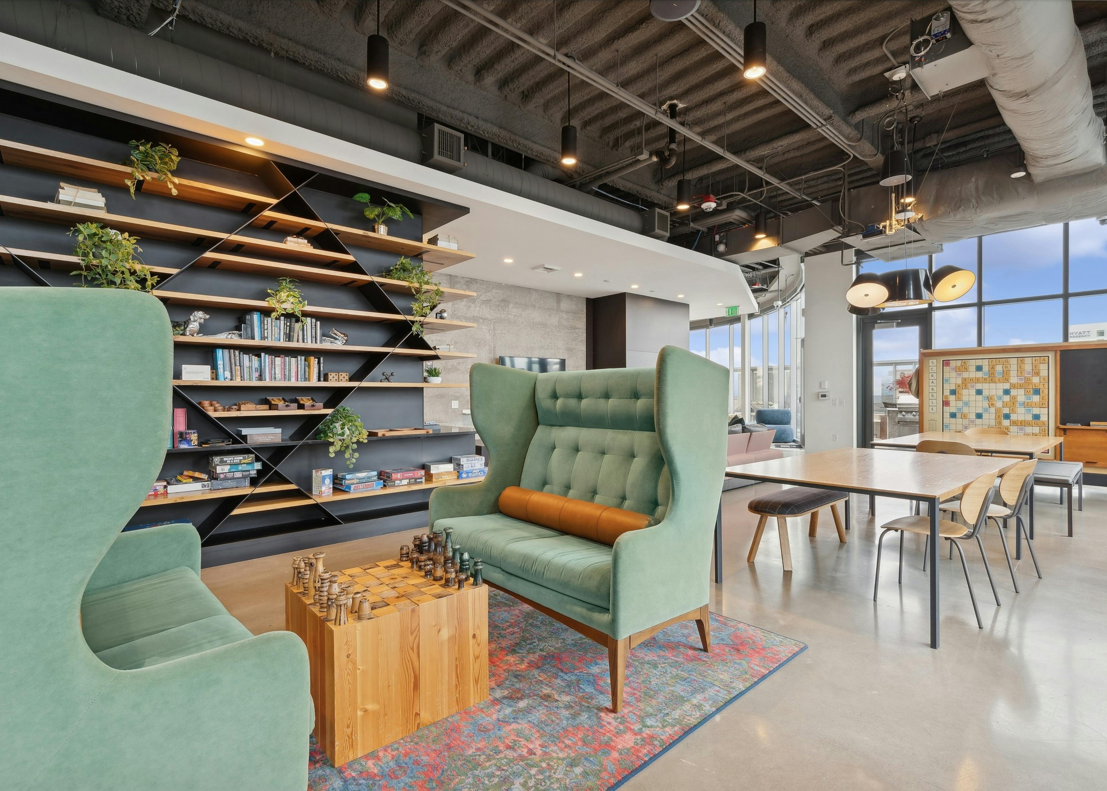 Game zone featuring green wingback chairs, a chessboard, wall shelving with books and games and a communal table at AMLI Arc