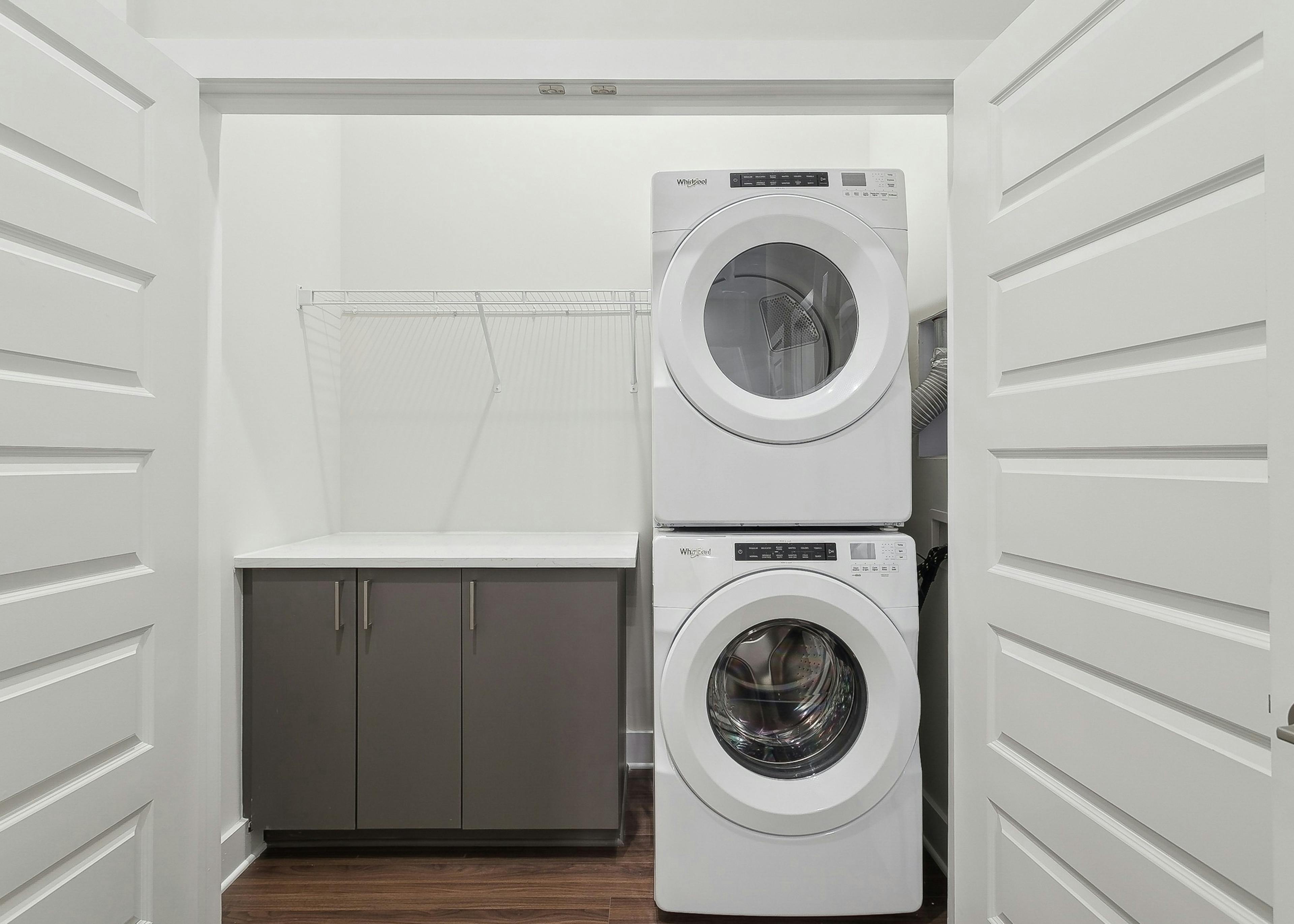 AMLI Brookhaven Laundry closet with full size stackable white washer and dryer beside a gray cabinet with shelf above it