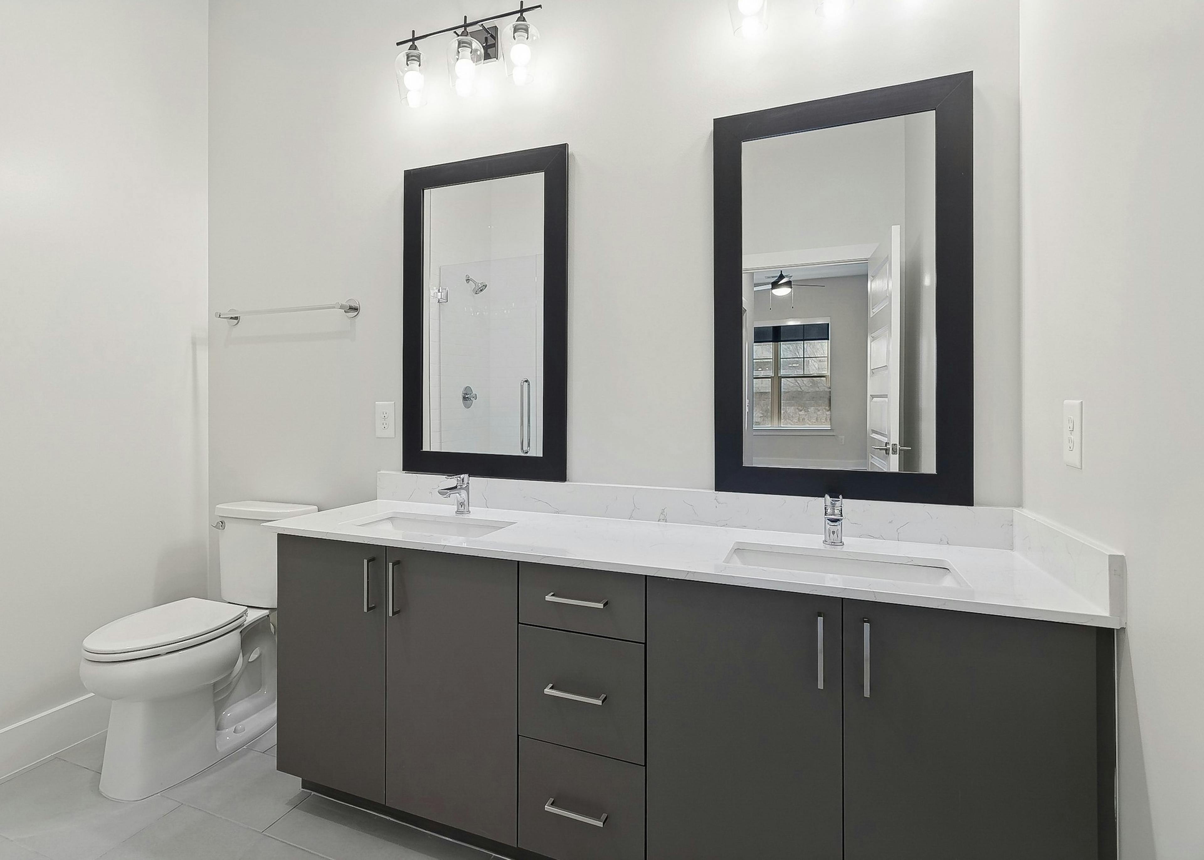 Bathroom at AMLI Brookhaven apartments with 2 large mirrors above a dark gray double vanity with white quartz countertop next to a toilet
