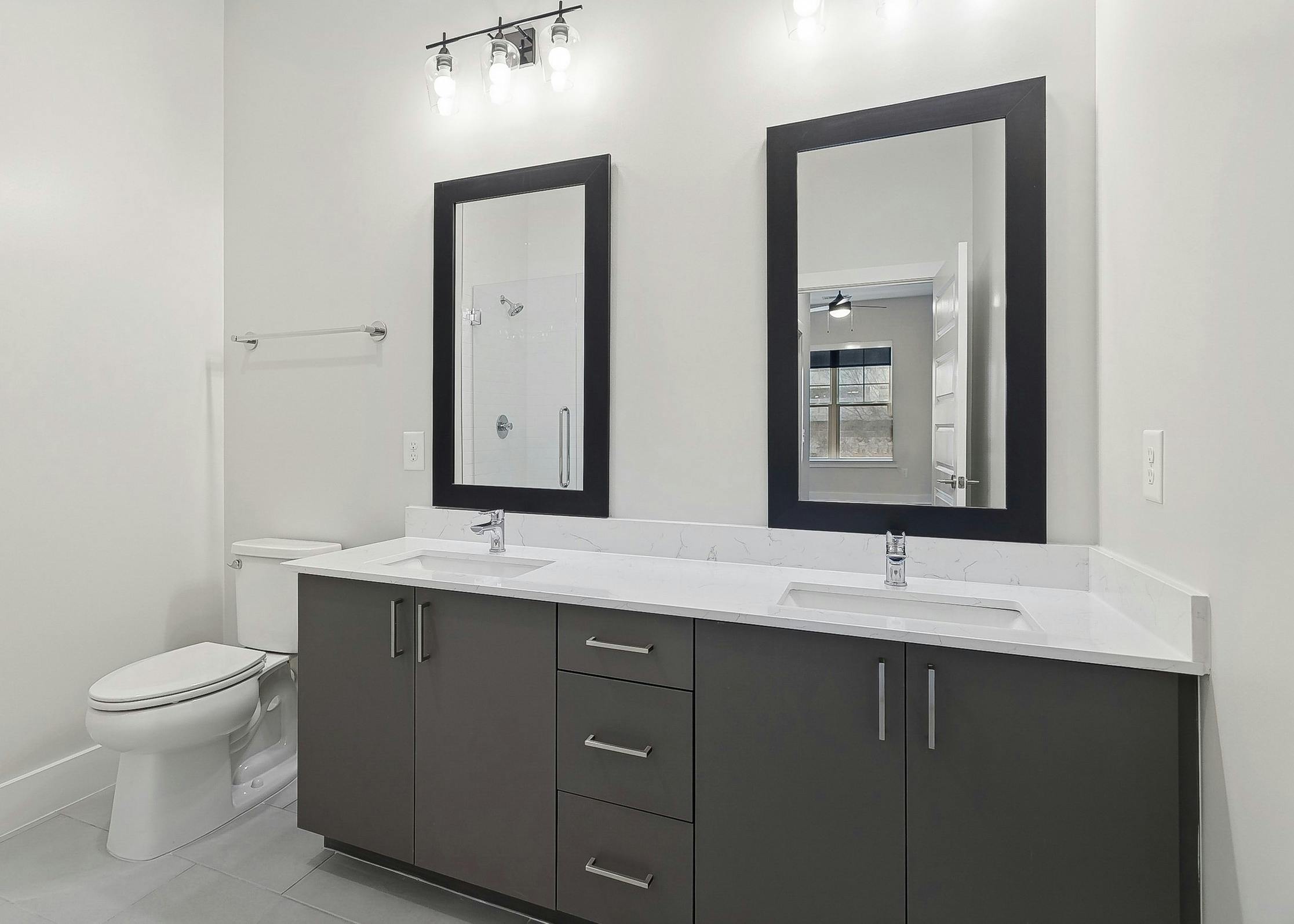 Bathroom at AMLI Brookhaven apartments with 2 large mirrors above a dark gray double vanity with white quartz countertop next to a toilet