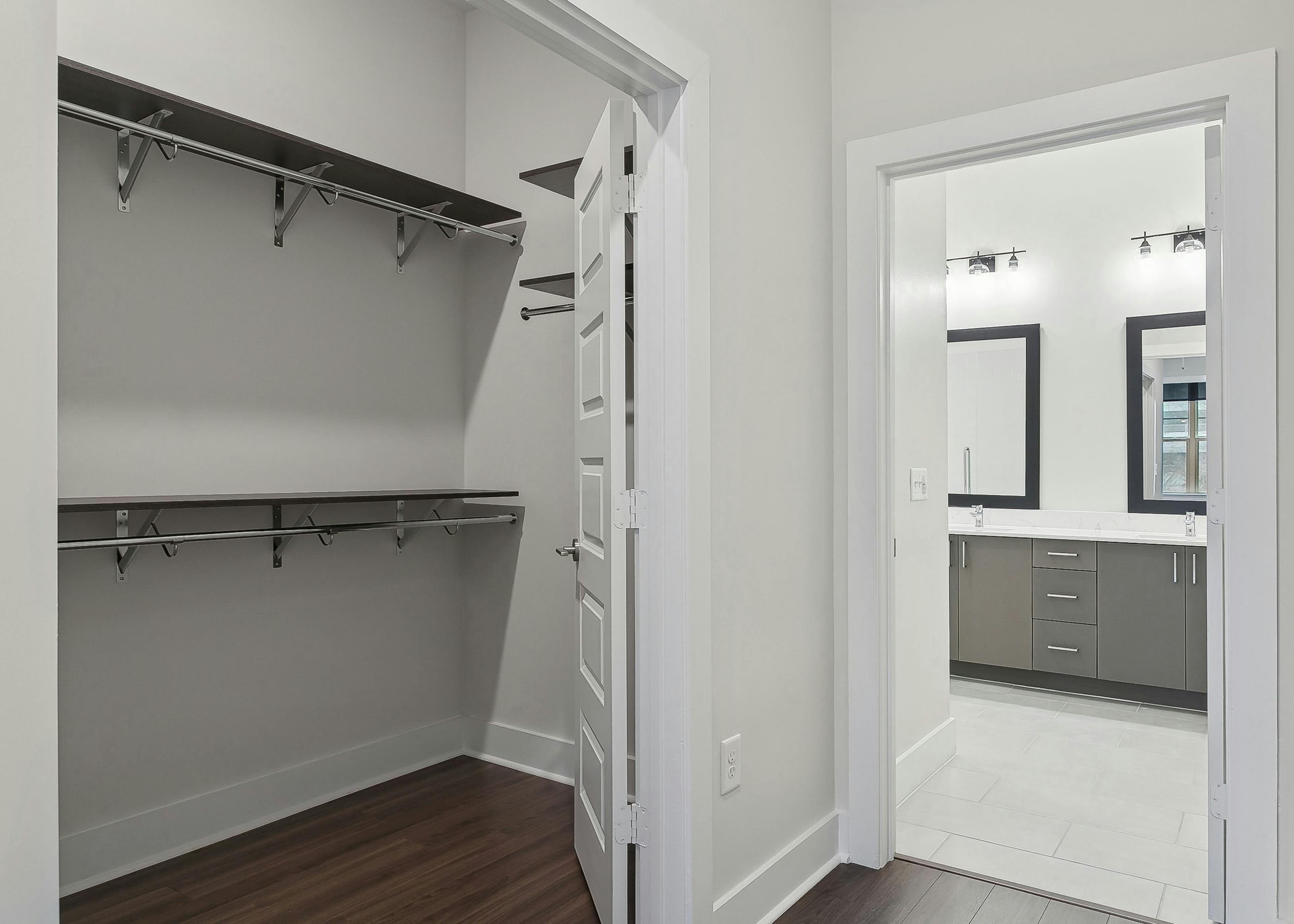 A walk in closet at AMLI Brookhaven apartments with wooden shelves and hanging rods with a view into a bathroom area with dual vanities