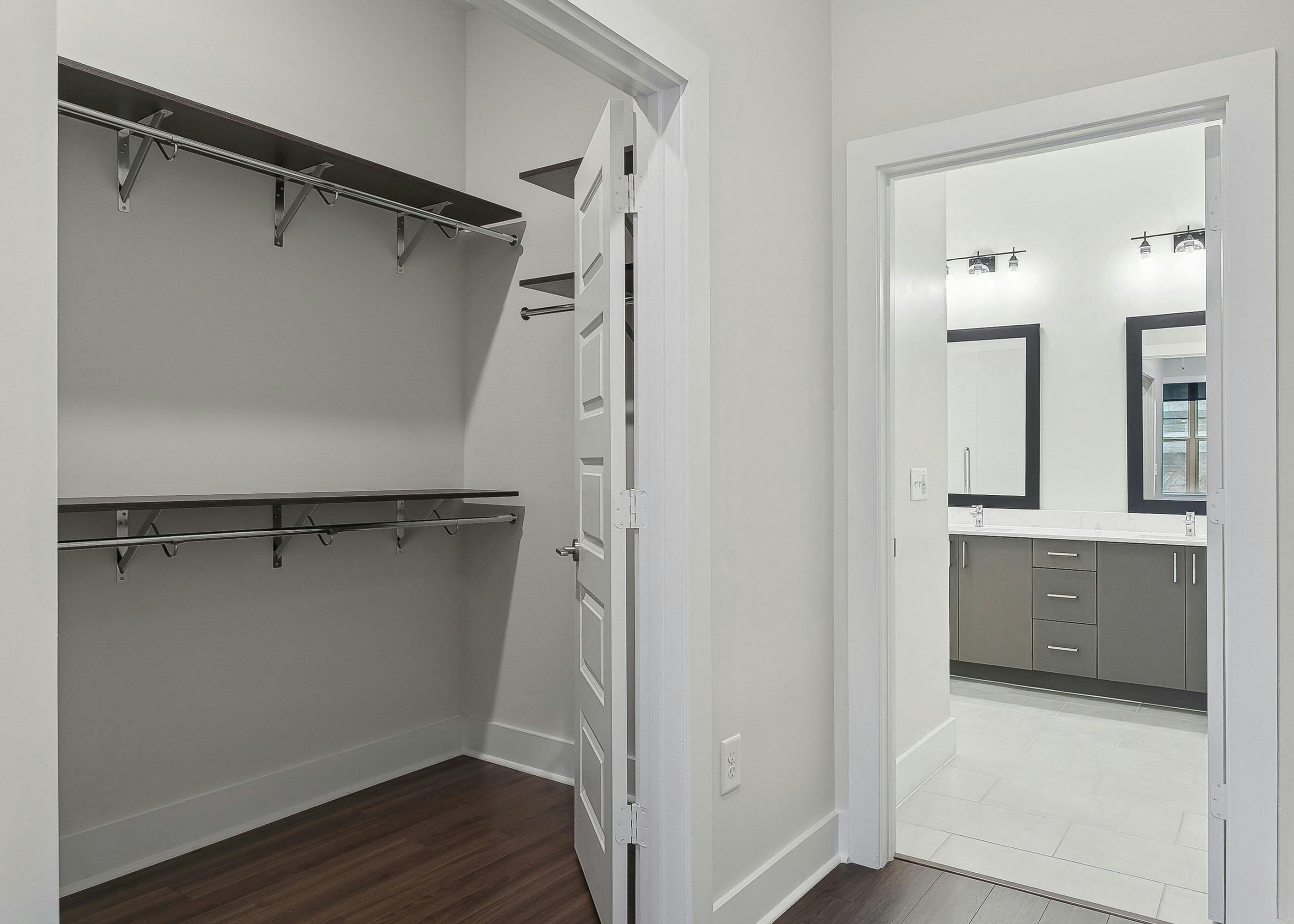 A walk in closet at AMLI Brookhaven apartments with wooden shelves and hanging rods with a view into a bathroom area with dual vanities