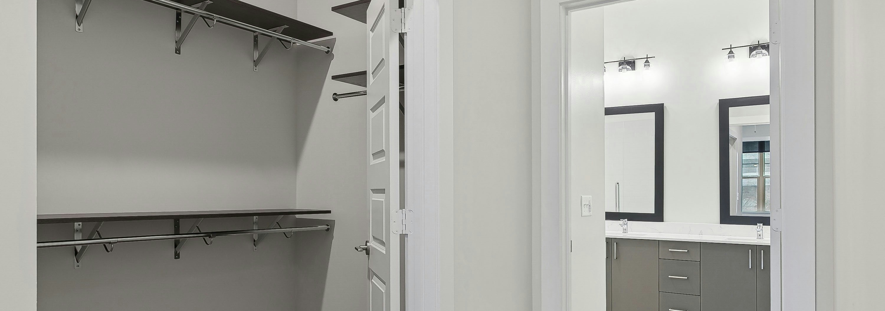A walk in closet at AMLI Brookhaven apartments with wooden shelves and hanging rods with a view into a bathroom area with dual vanities
