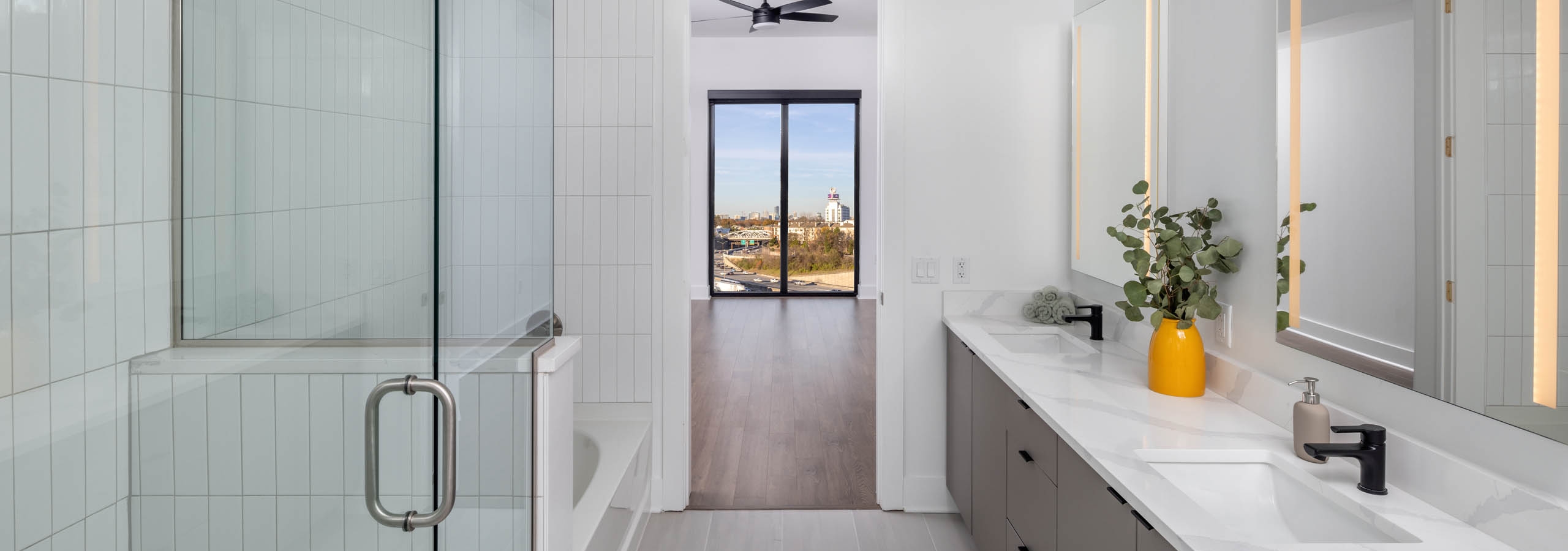 Modern bathroom with grey tile, a glass shower, bathtub and double vanity at AMLI Atlantic Station looking onto a bedroom with a city view.