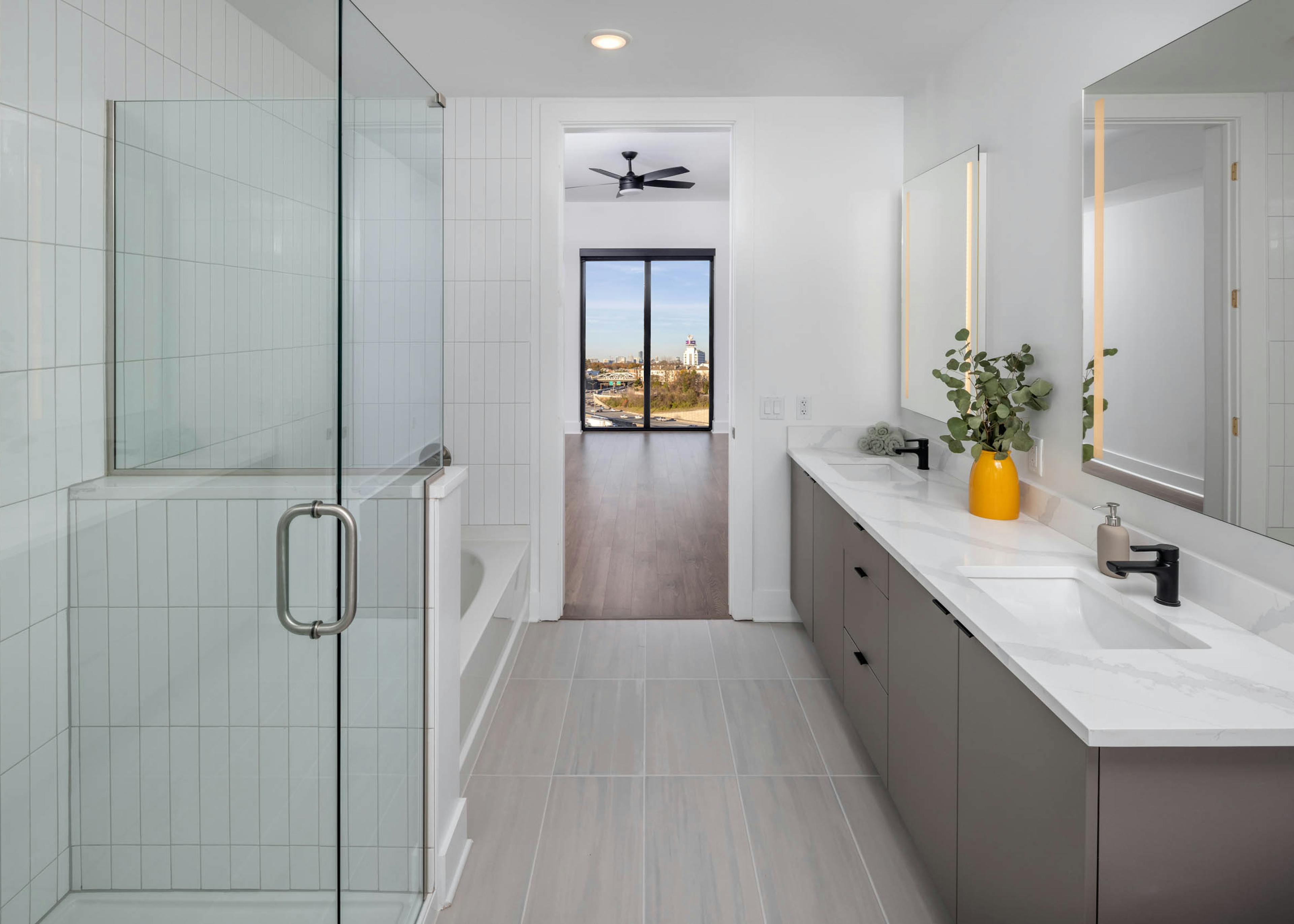 Modern bathroom with grey tile, a glass shower, bathtub and double vanity at AMLI Atlantic Station looking onto a bedroom with a city view.