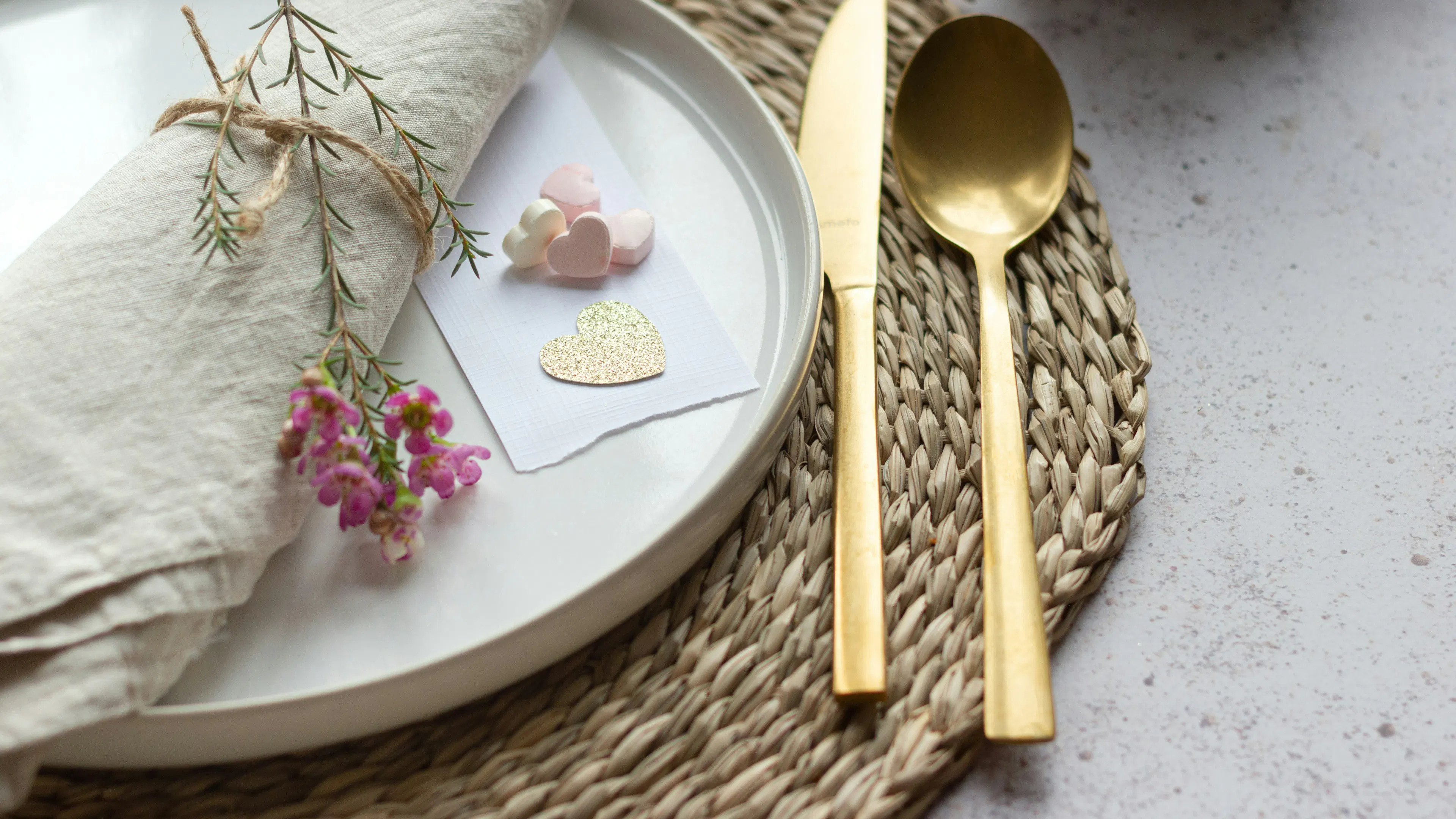 A white plate on a rattan tablecloth next to gold cutlery and a few dried flowers for decoration