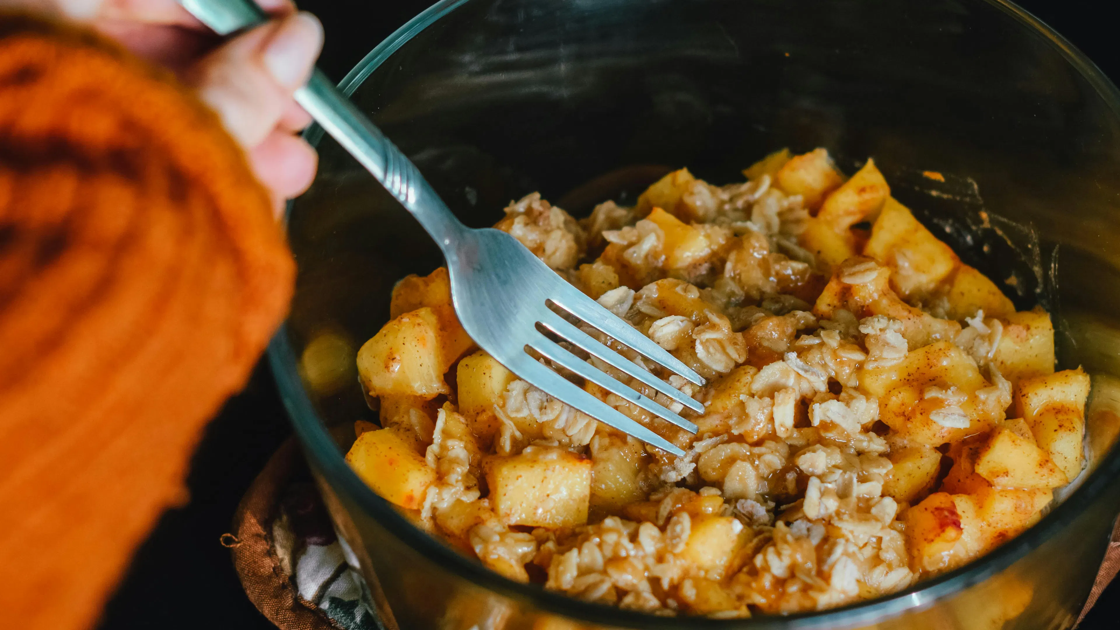 A bowl of peach cobbler mix in a glass bowl with a woman's hand mixing it with a fork