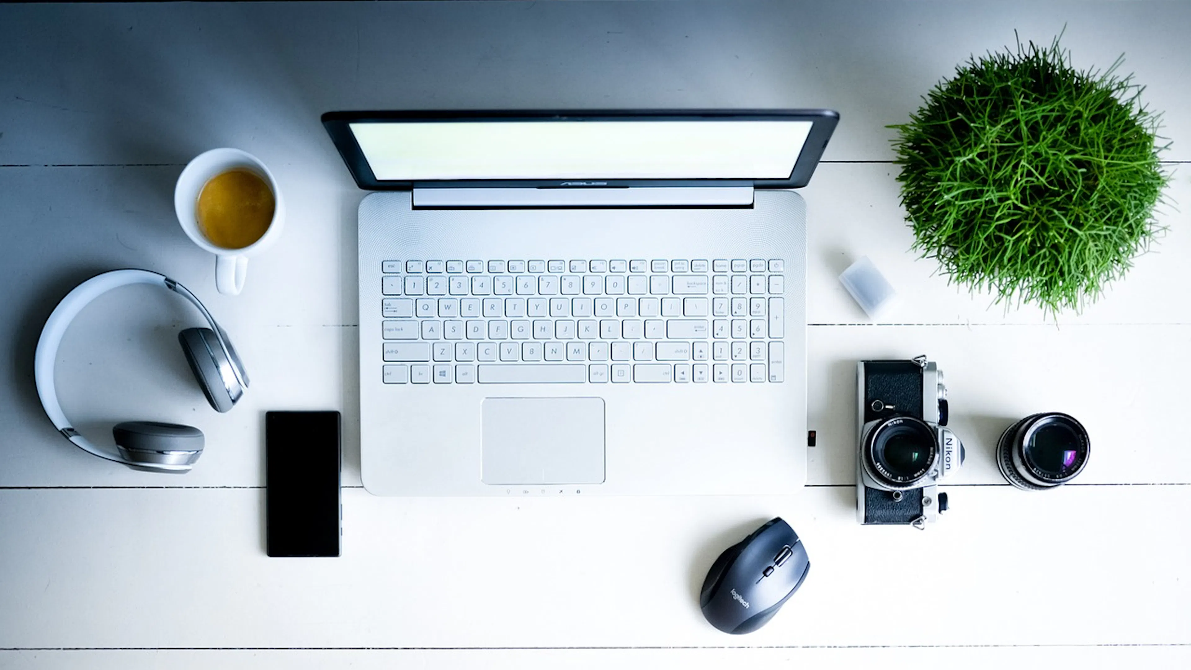 white desk with open lap top and green plant and headphones and coffee and camera