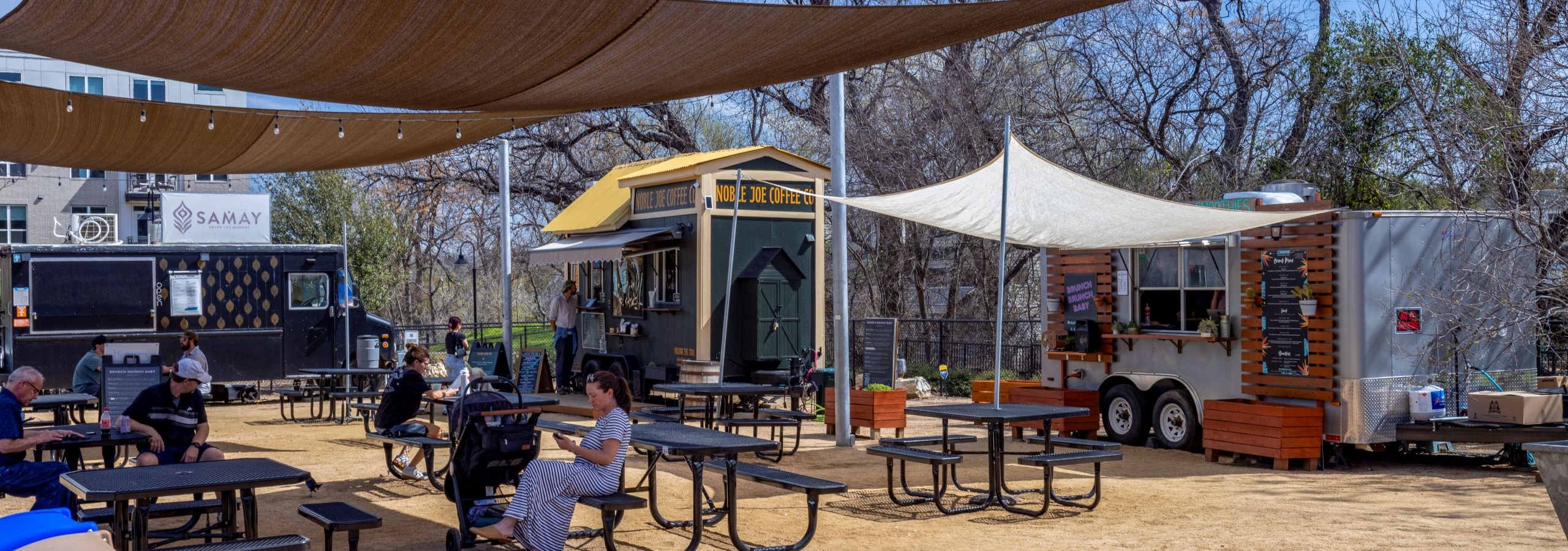AMLI South Shore resident food truck event set on tan gravel with people sitting at picnic tables under shade surrounded by 3 food trucks
