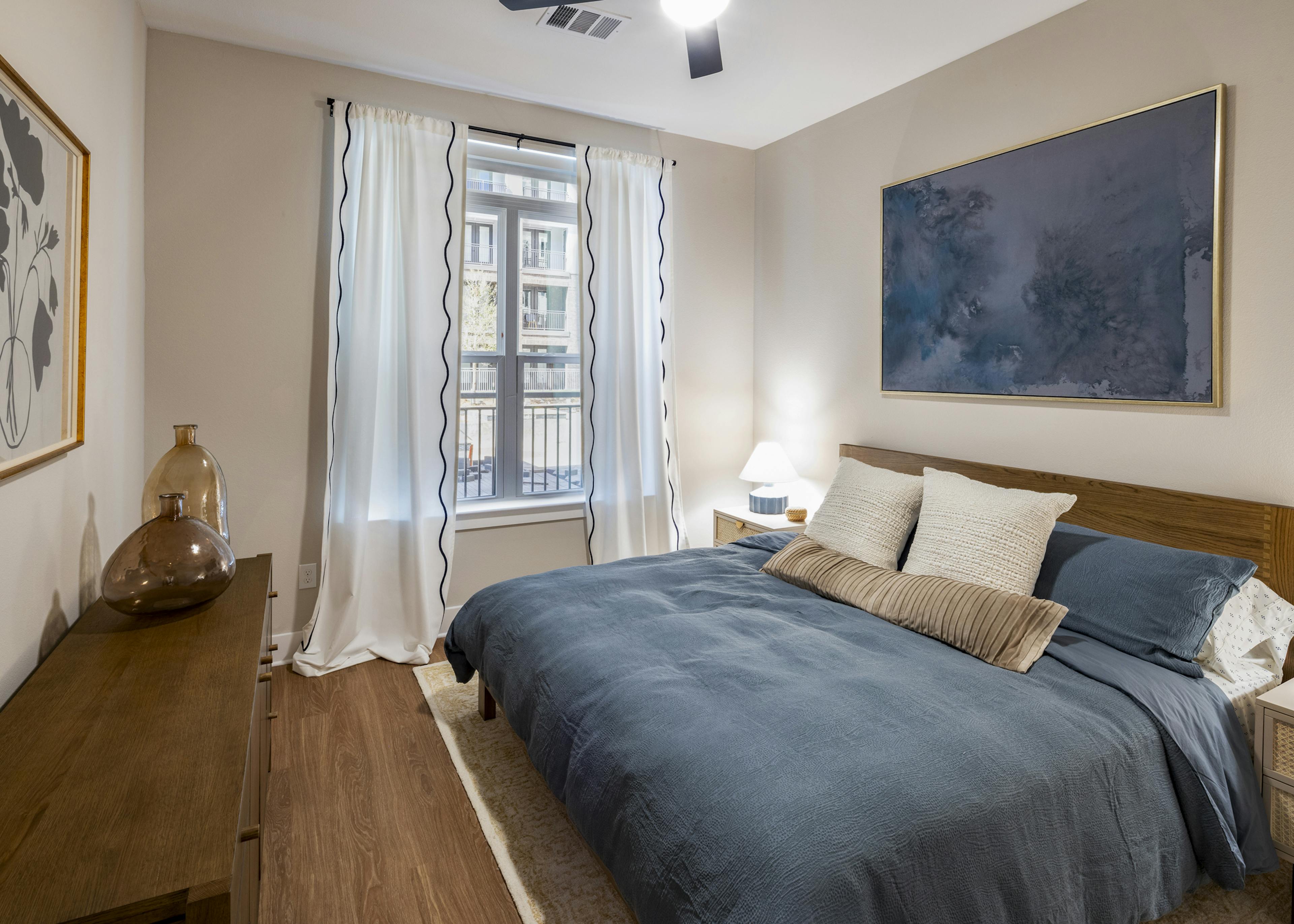 Interior view of a bedroom at AMLI North Park apartments with a wooden bed frame, blue bedding, large window, and abstract wall art