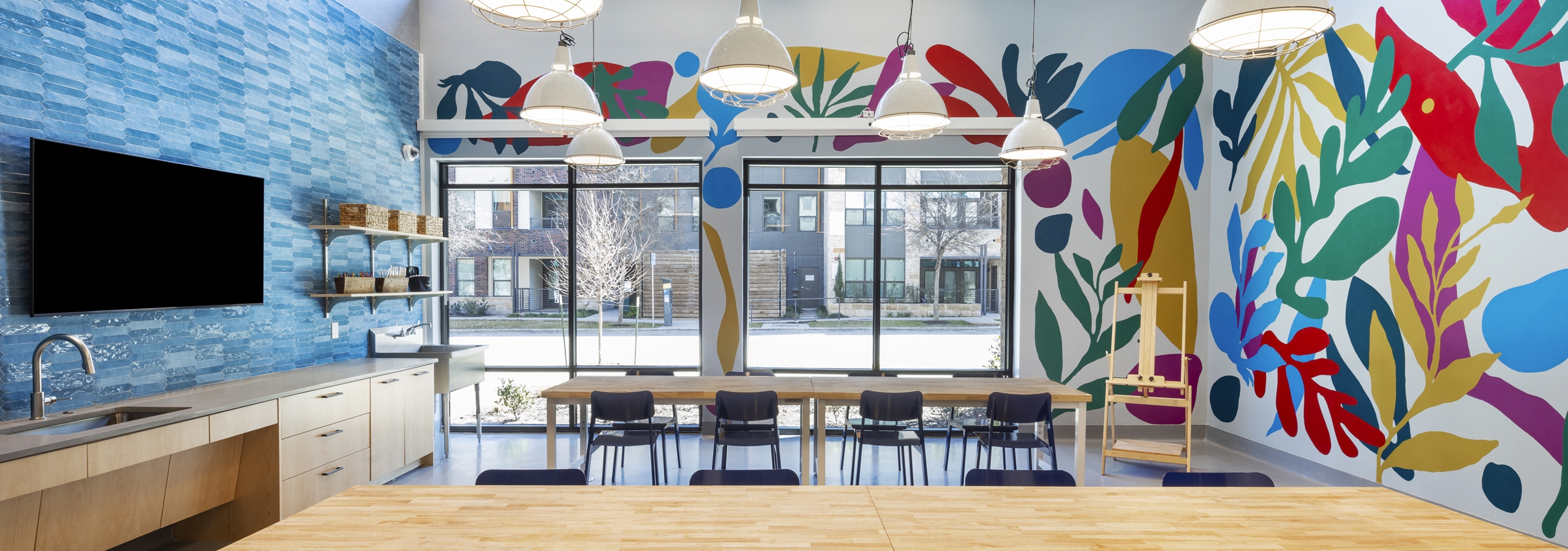Creative studio at AMLI North Park with colorful murals, large tables, pendant lighting, and a TV mounted on blue tile backsplash wall
