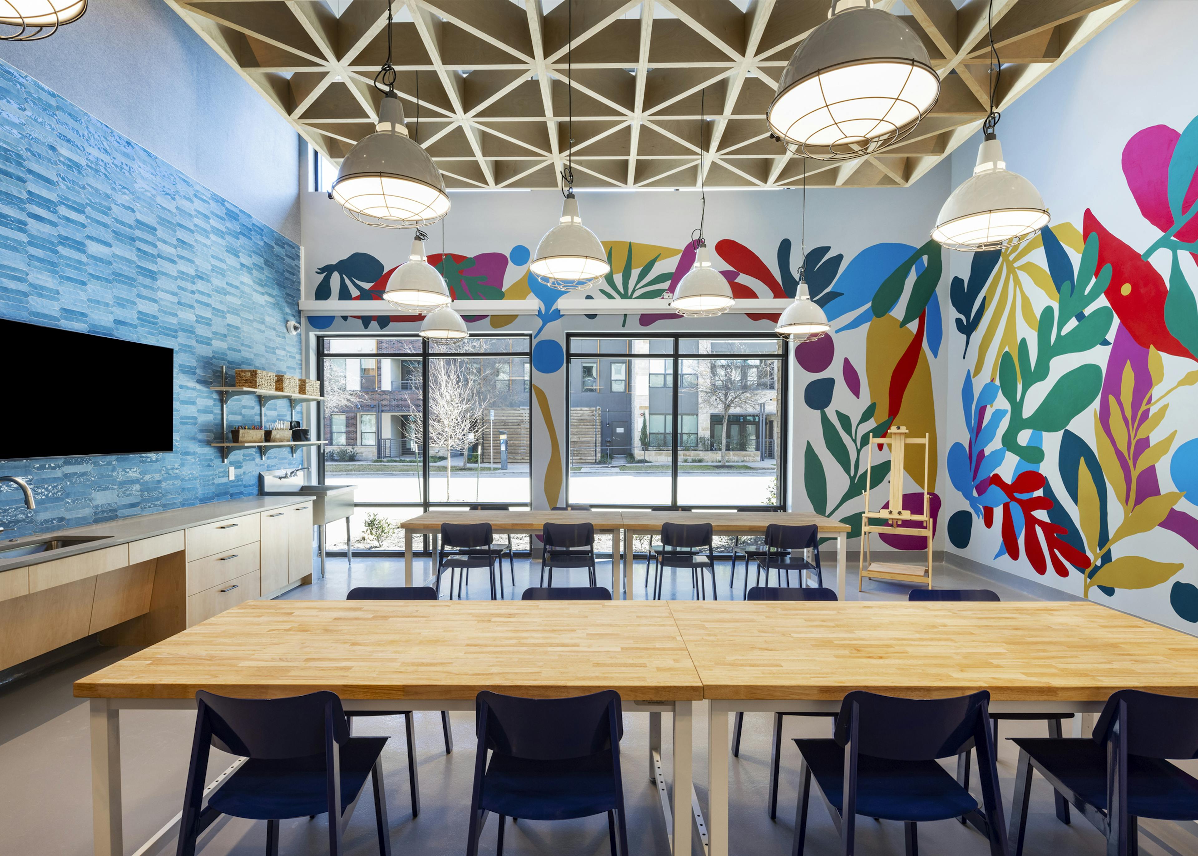 Creative studio at AMLI North Park with colorful murals, large tables, pendant lighting, and a TV mounted on blue tile backsplash wall