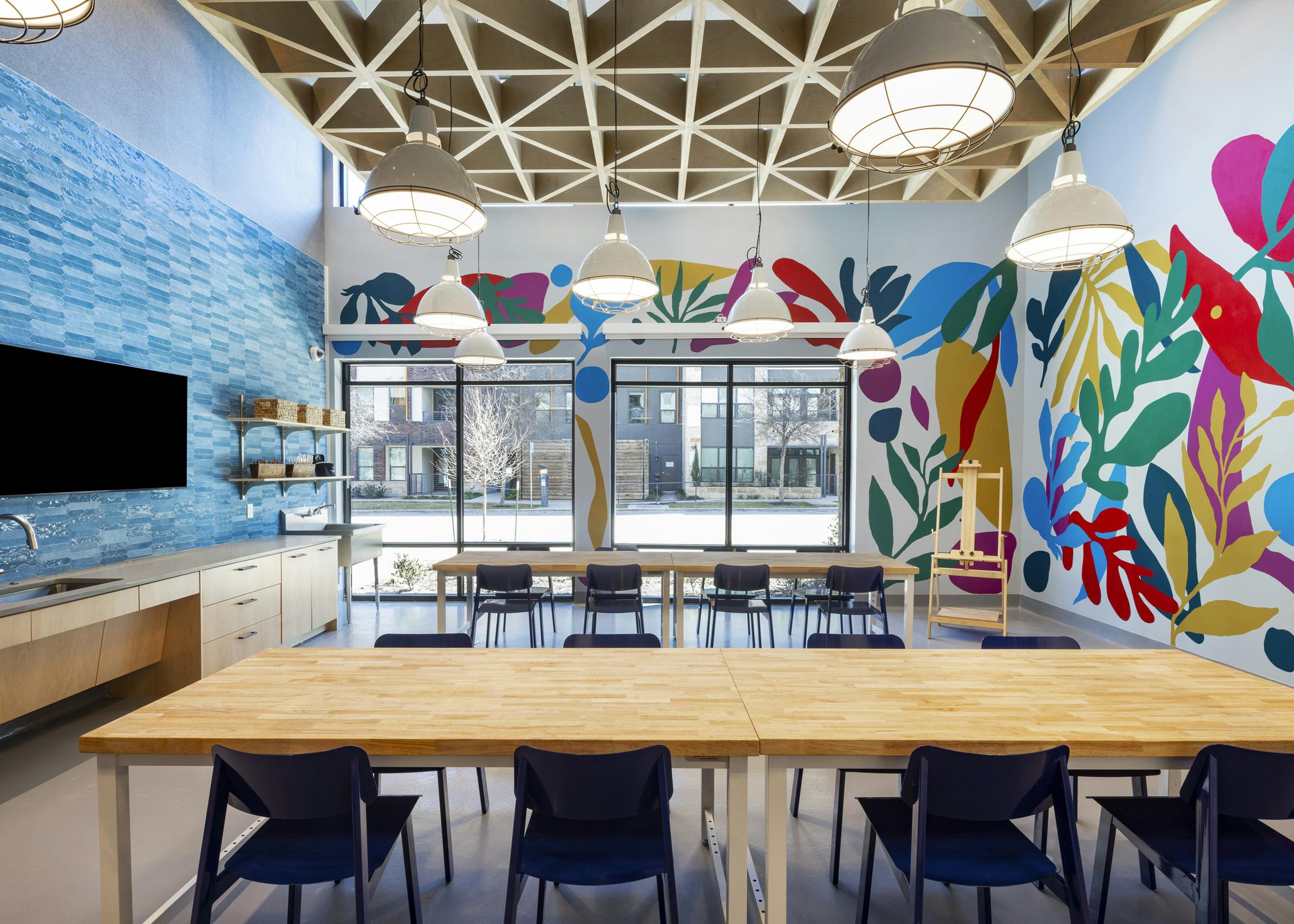 Creative studio at AMLI North Park with colorful murals, large tables, pendant lighting, and a TV mounted on blue tile backsplash wall