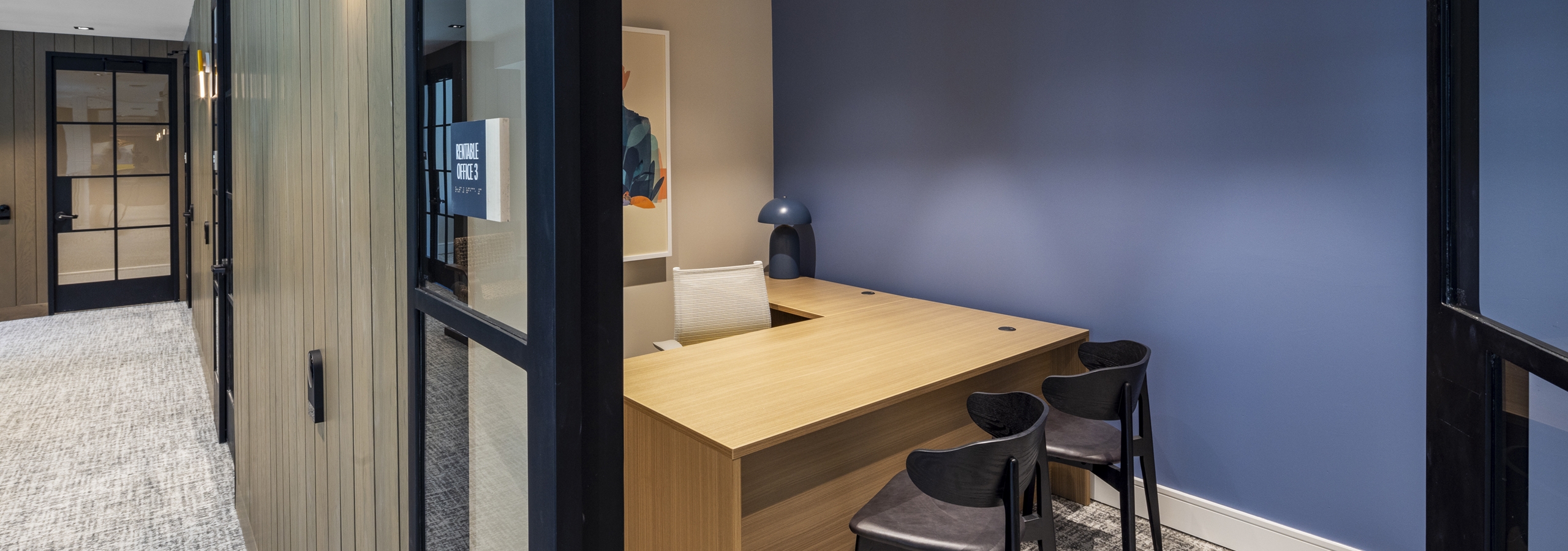 Private office at AMLI North Park featuring a wood desk, black chairs, a blue accent wall, and glass doors for a quiet workspace