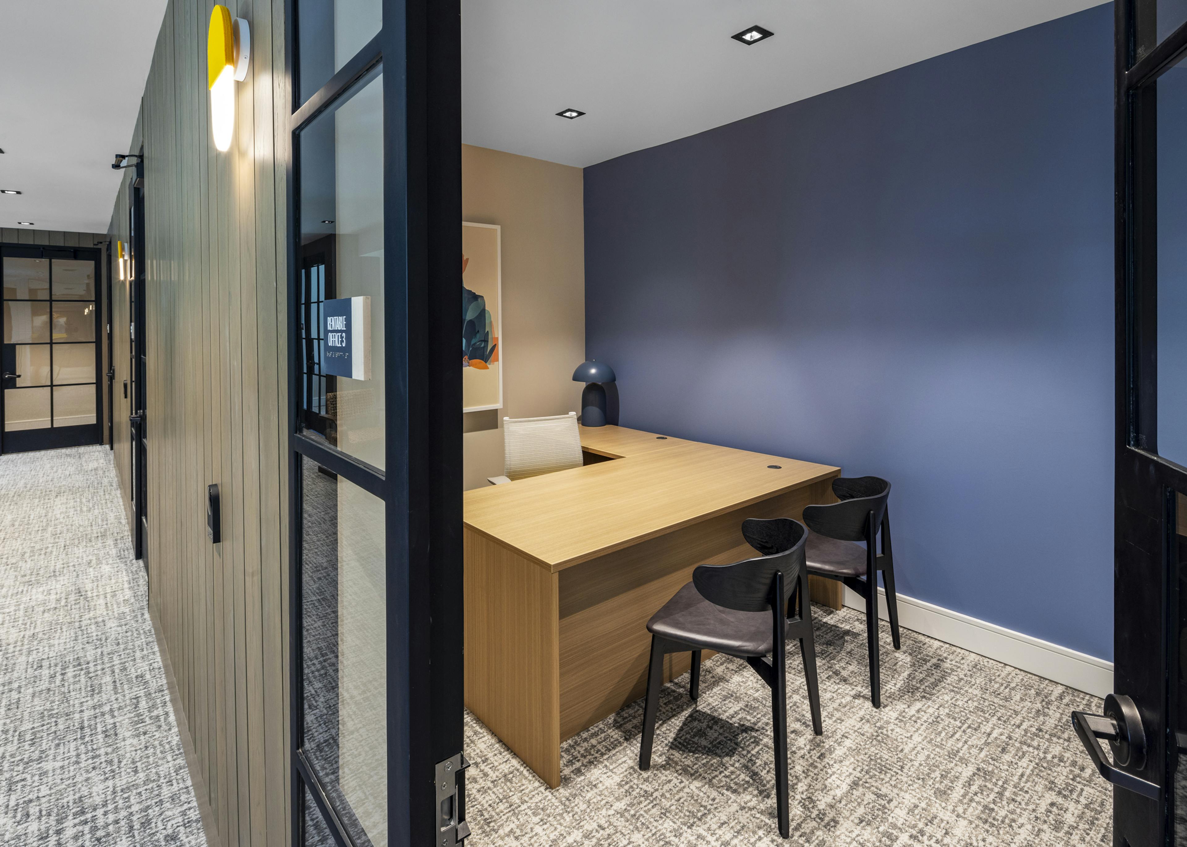 Private office at AMLI North Park featuring a wood desk, black chairs, a blue accent wall, and glass doors for a quiet workspace