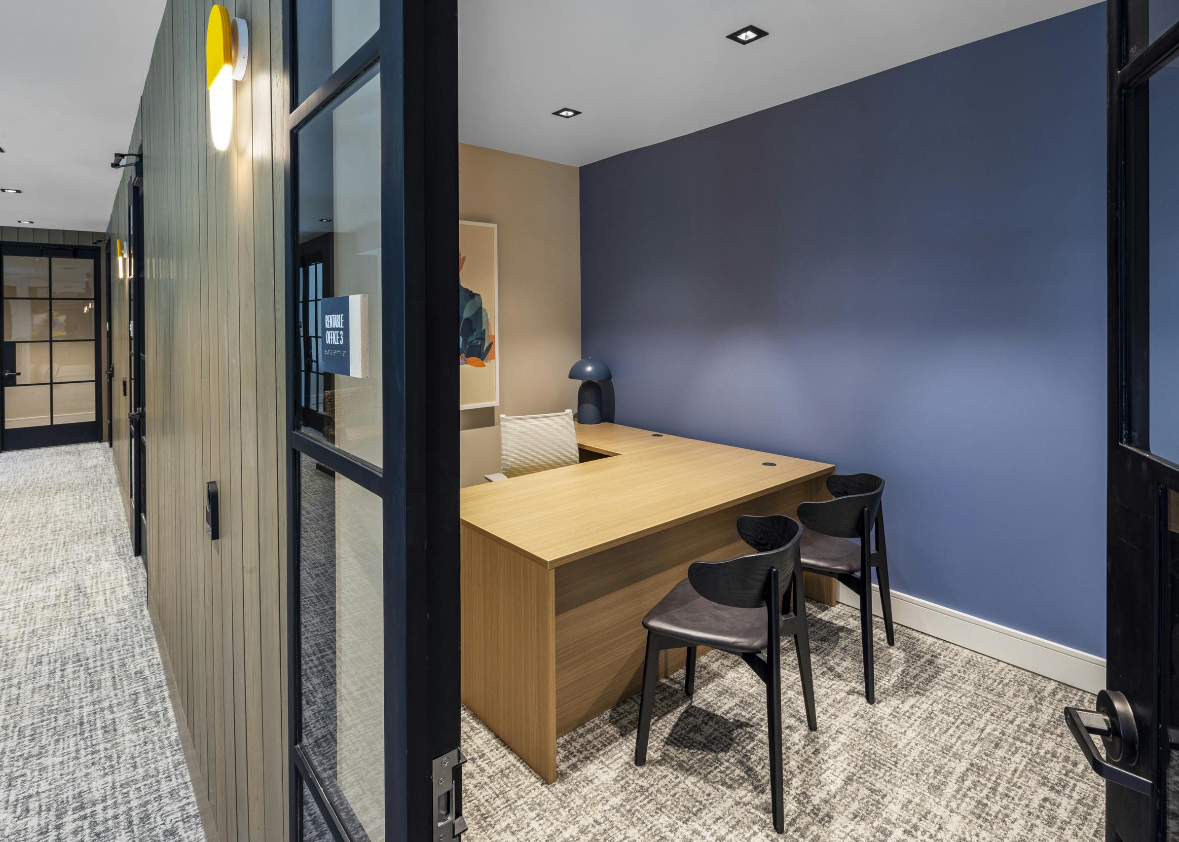 Private office at AMLI North Park featuring a wood desk, black chairs, a blue accent wall, and glass doors for a quiet workspace