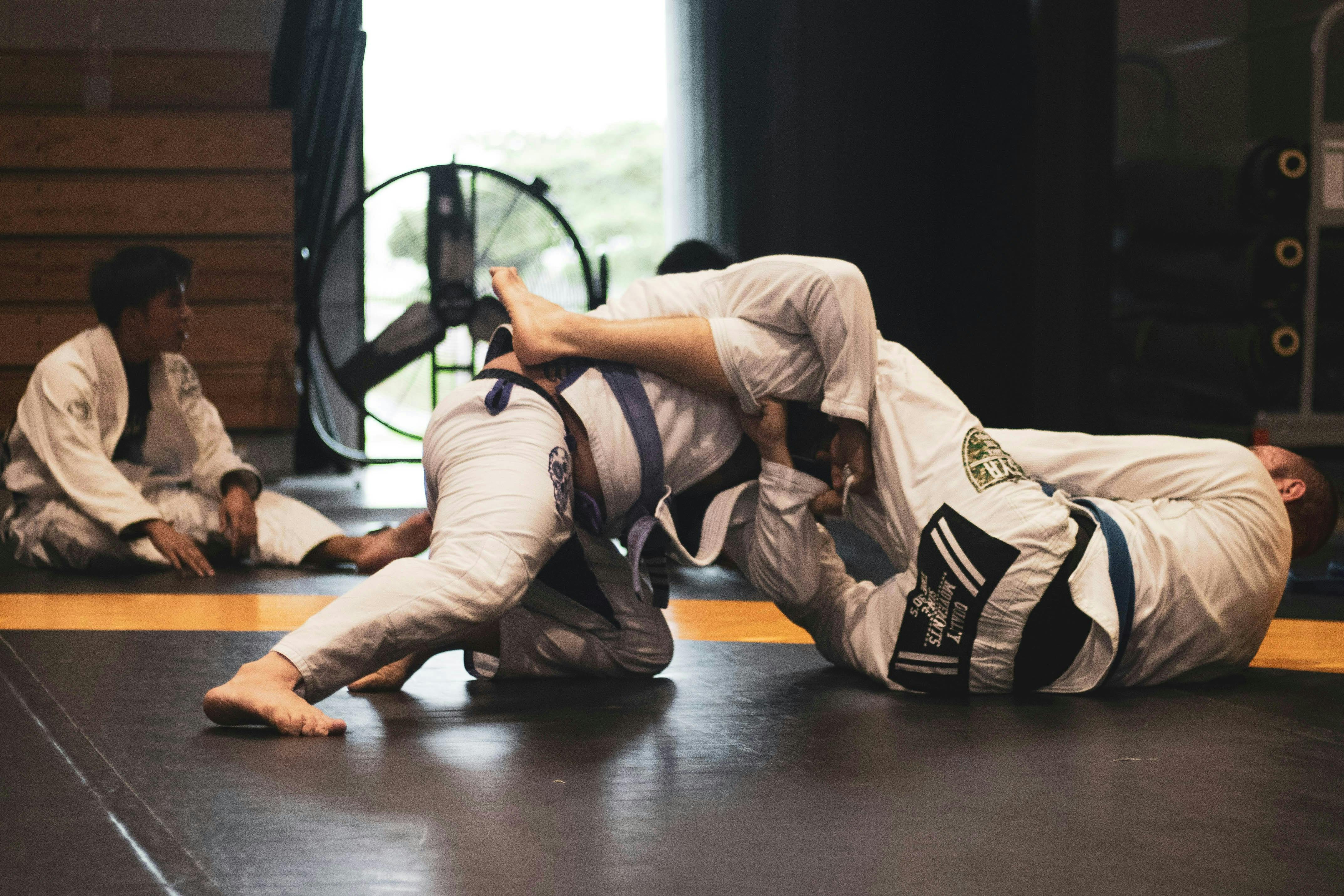 Two grapplers in Jiu-Jitsu uniforms sparring on a black floor mat