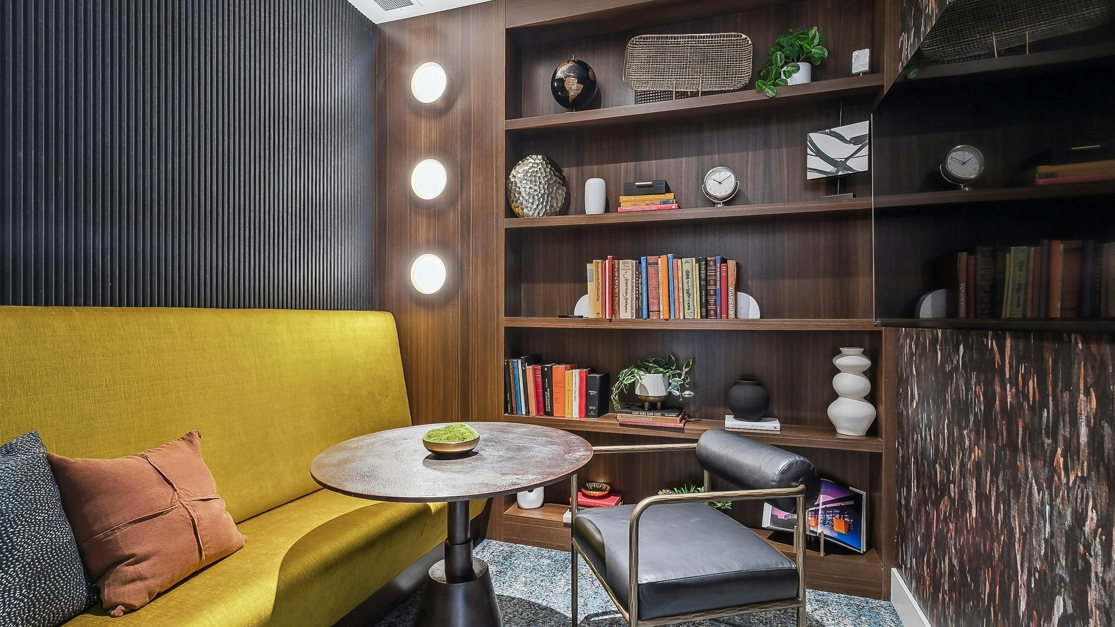A cozy resident lounge with a mustard yellow couch and a round wooden table with a black chair and shelving with books