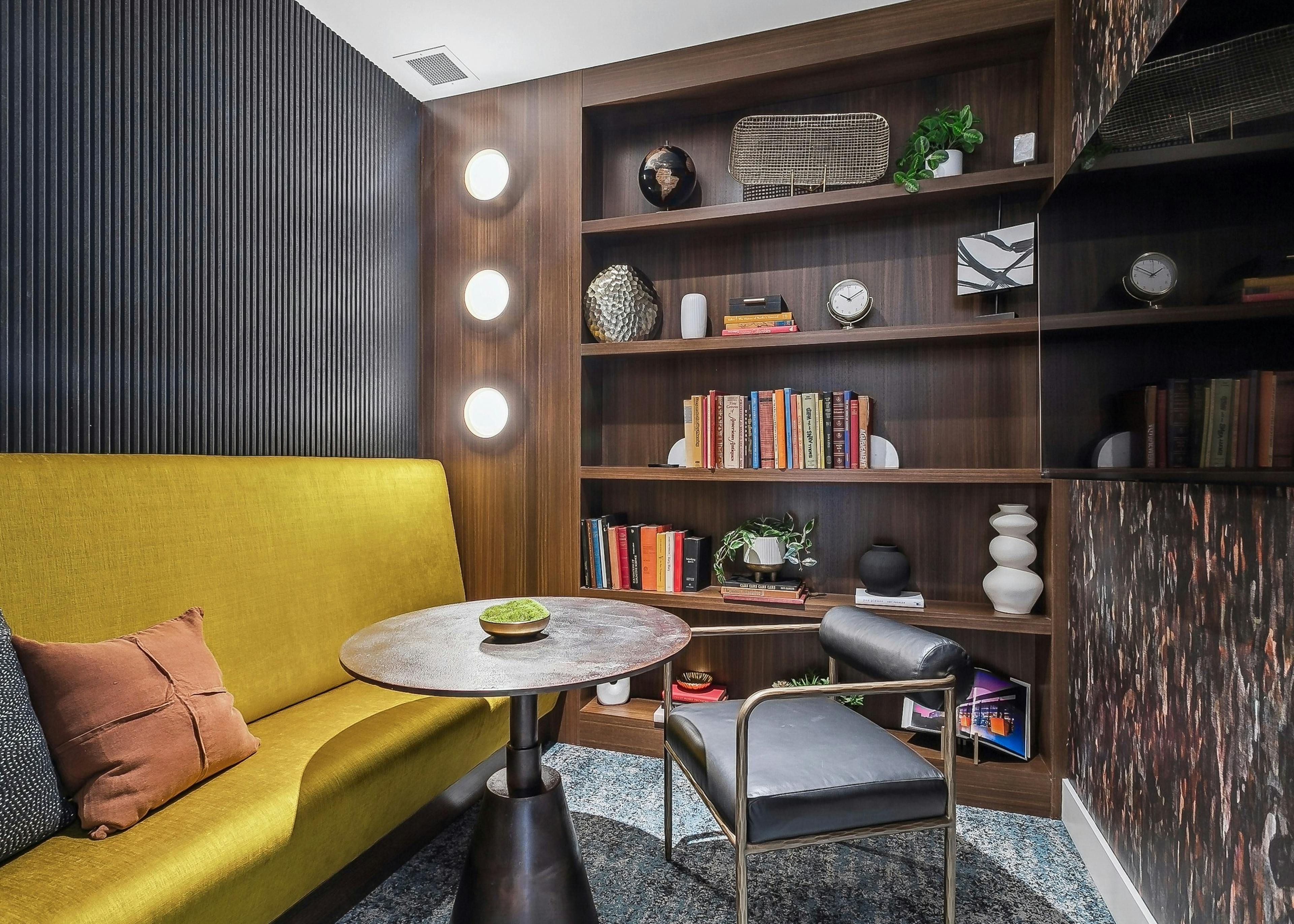 A cozy resident lounge with a mustard yellow couch and a round wooden table with a black chair and shelving with books