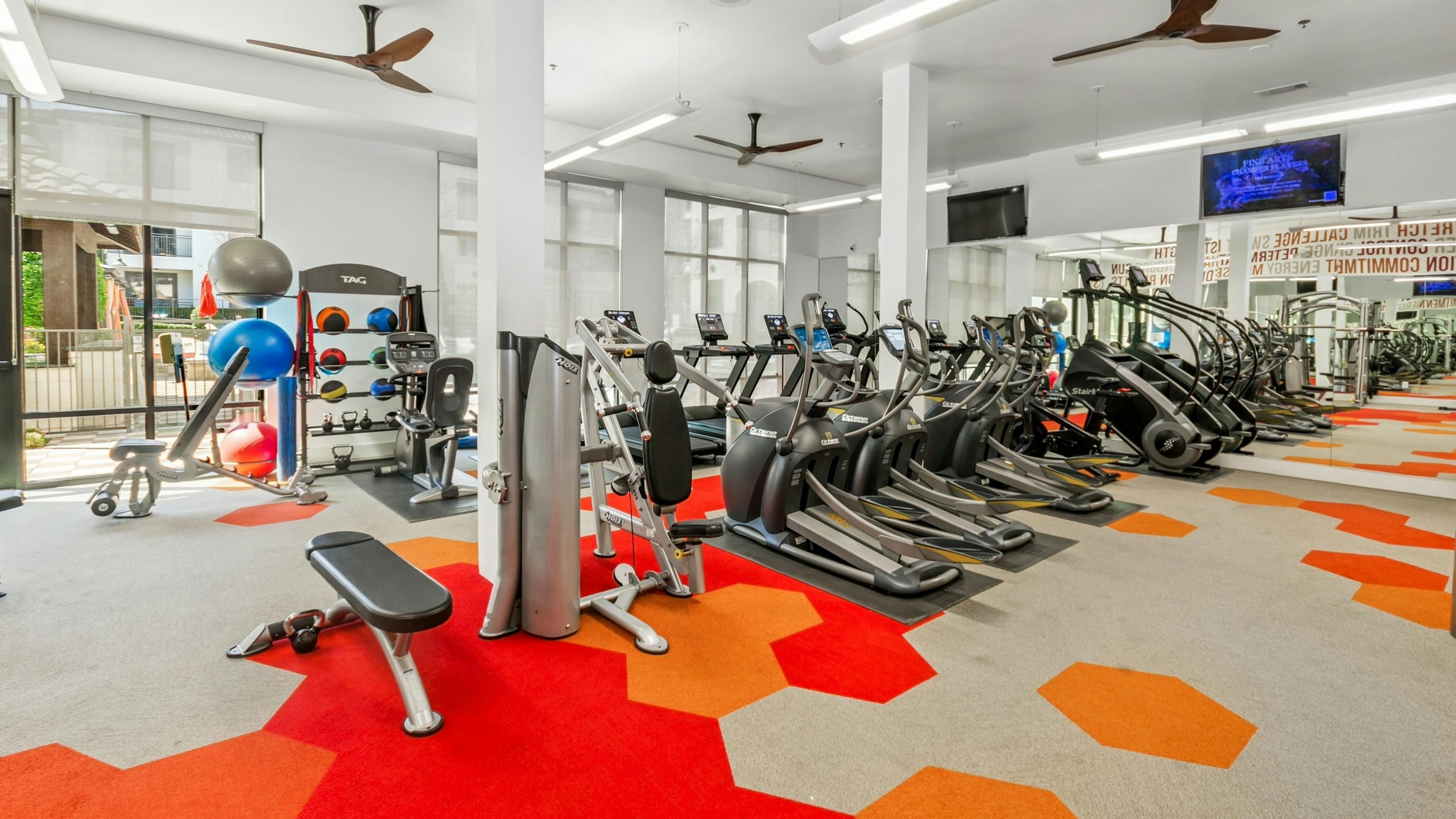 Modern gym at AMLI on Riverside with treadmills, ellipticals, and weight machines with a bright red and orange hexagonal patterned floor