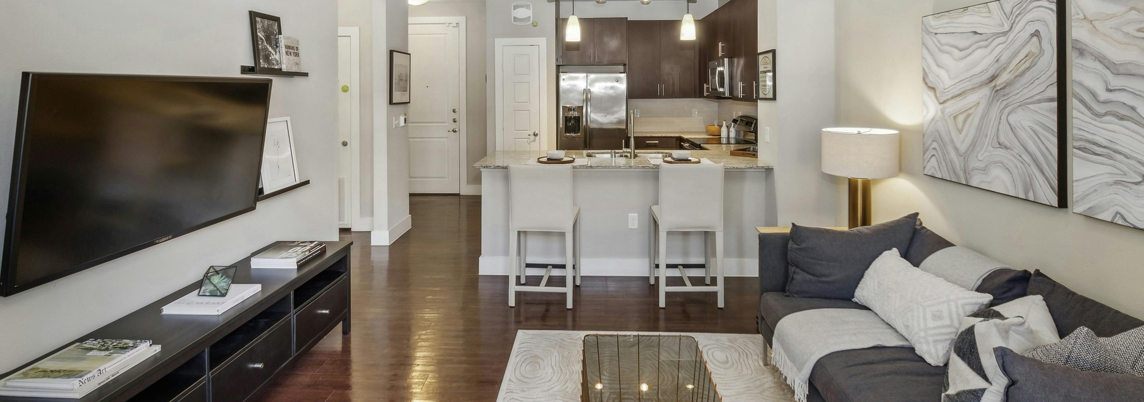 AMLI West Plano living room with gray couch facing TV on hardwood floors looking toward an open kitchen with breakfast bar 