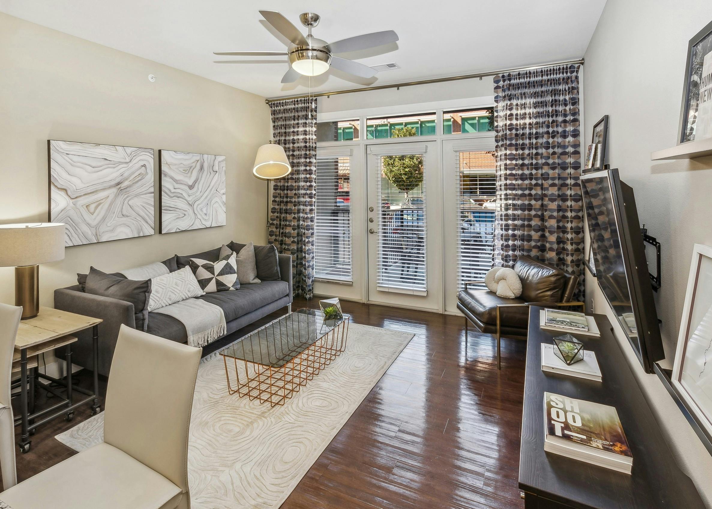 Apartment living room with gray couch on light rug on wood floors and decorative curtains around a patio door and windows at AMLI West Plano