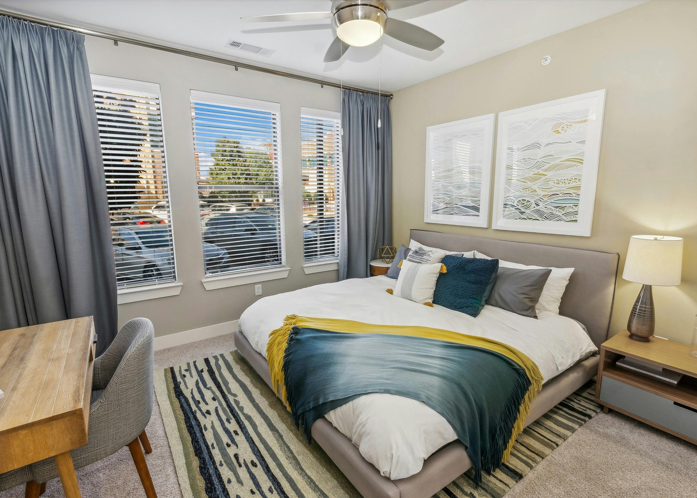 AMLI West Plano bedroom with king bed  with art above it and wood desk and three large windows with gray floor to ceiling curtains
