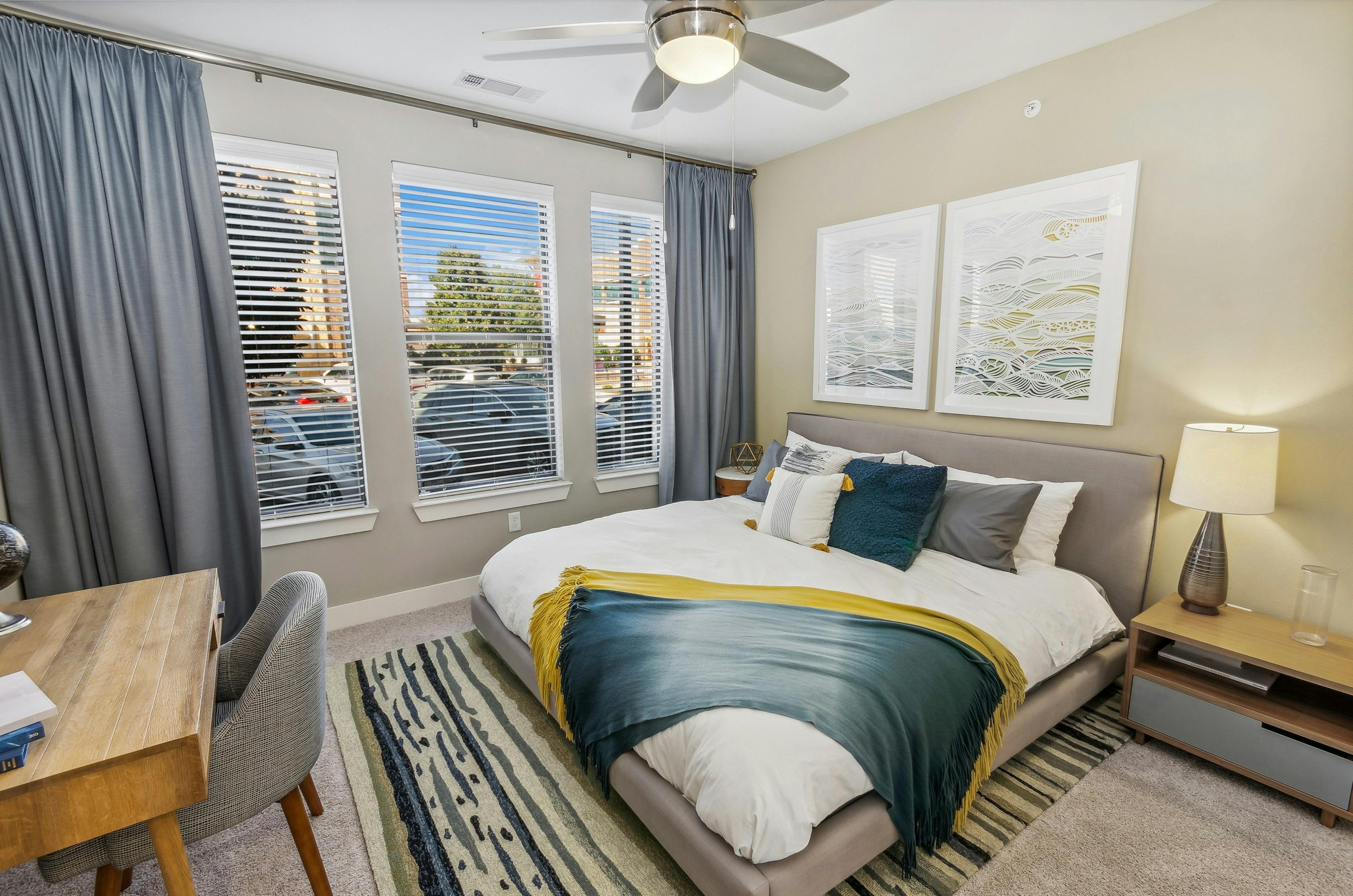 AMLI West Plano bedroom with king bed  with art above it and wood desk and three large windows with gray floor to ceiling curtains 