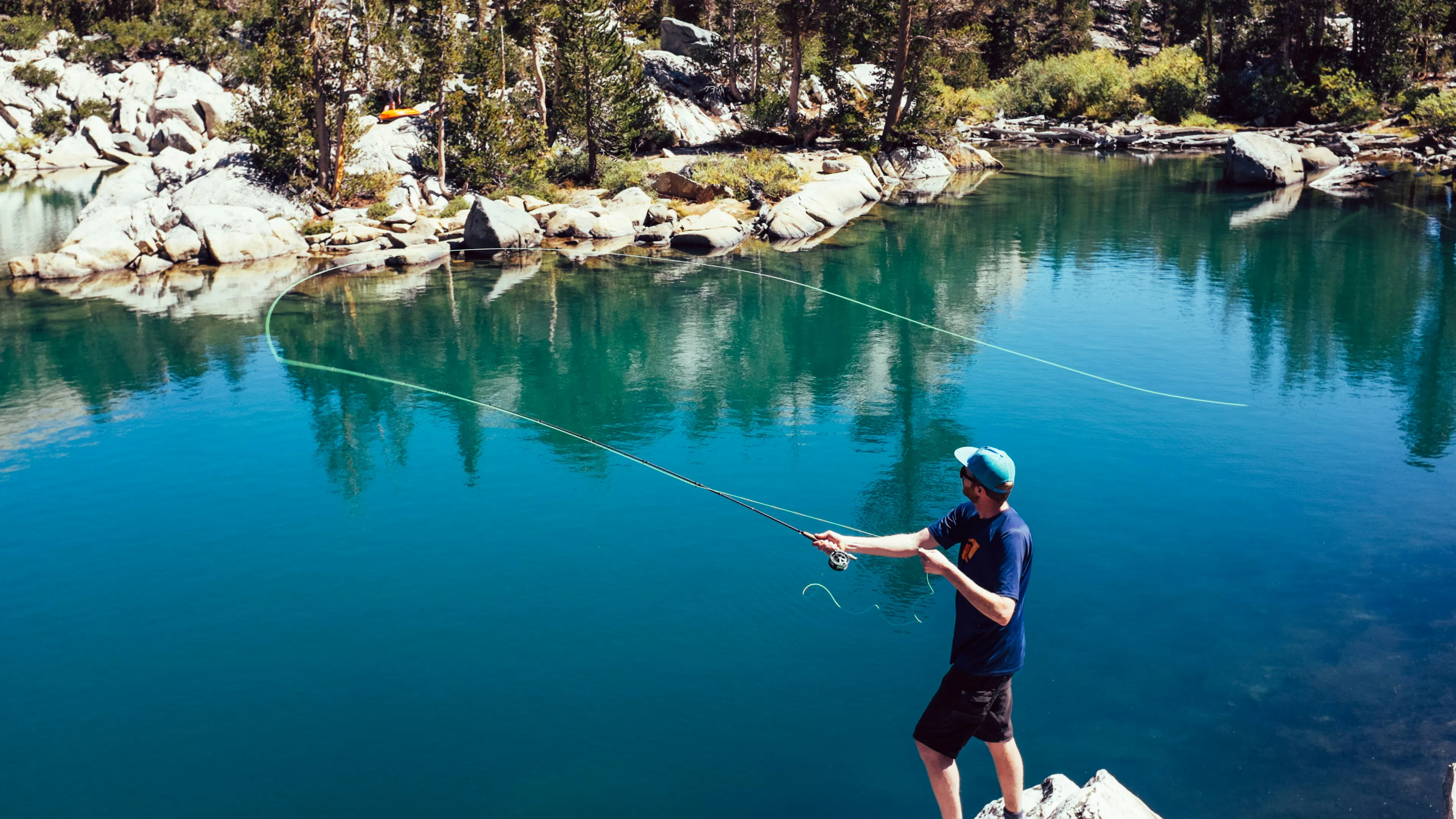 A man with a fly fishing rod casting into the clear blue waters of a rocky lined alpine lake