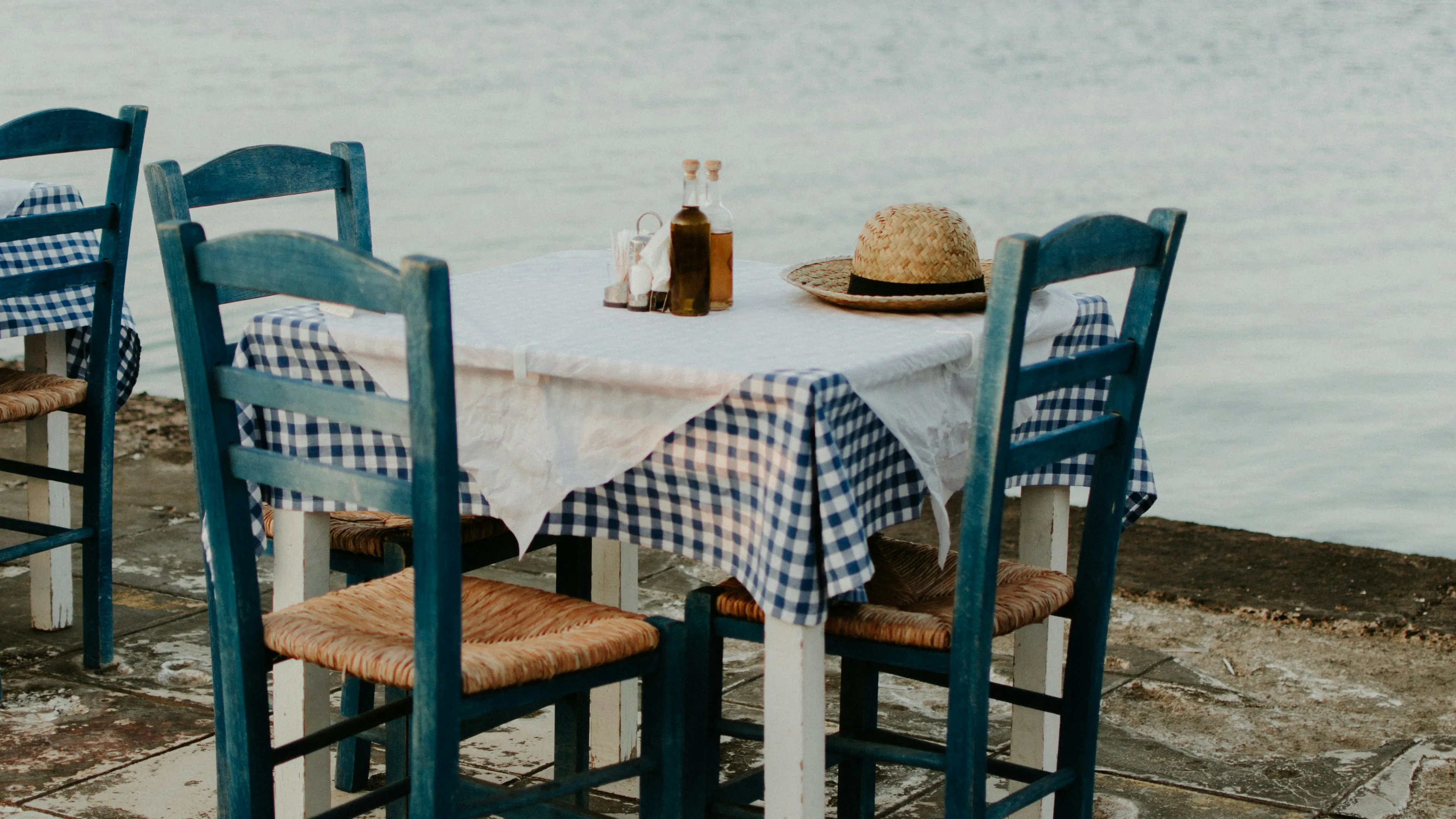 A small wooden table and three chairs with a blue tablecloth and some condiments overlooking the water