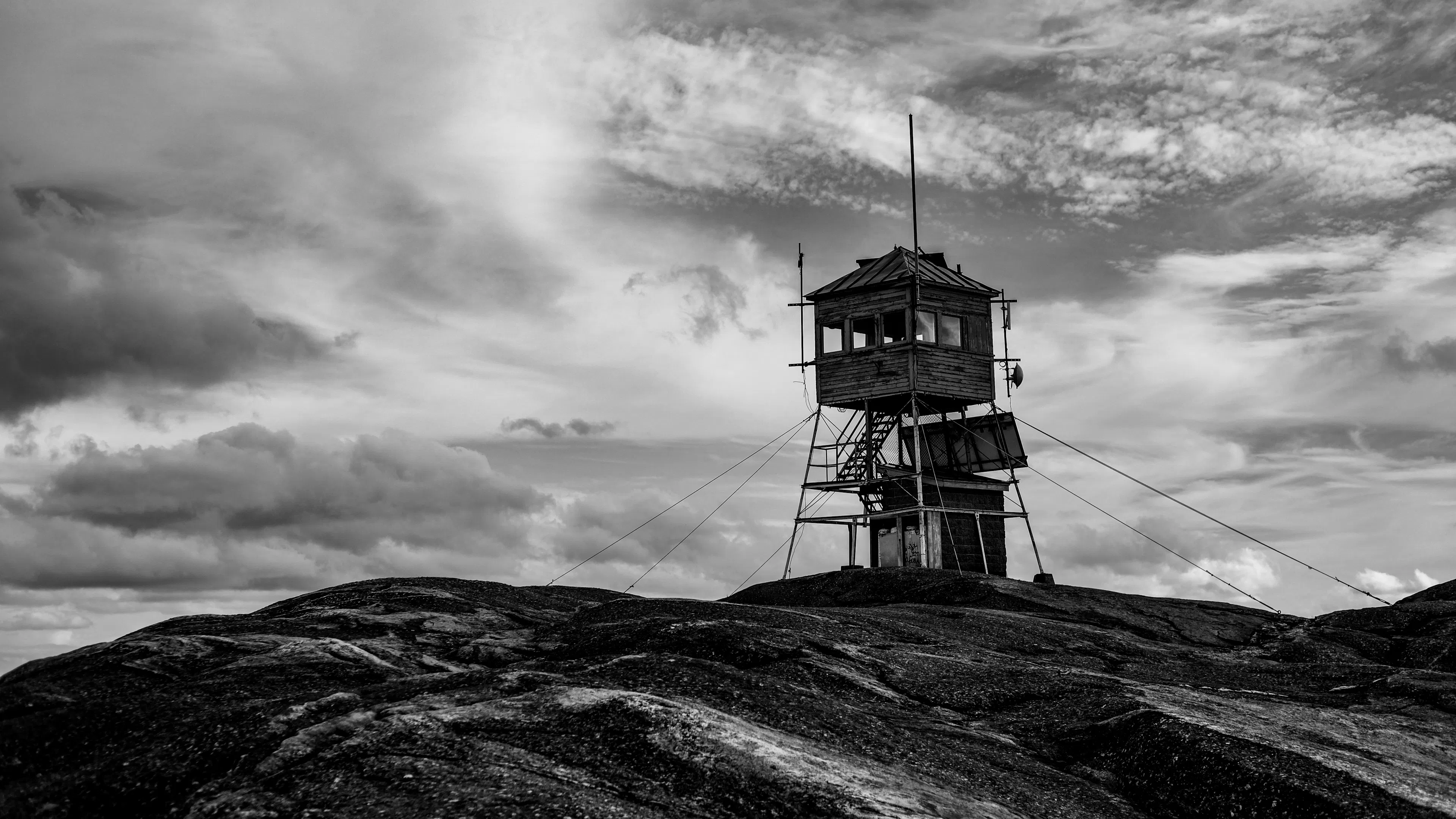A black and white view of a fire lookout on a bare peak silhouetted against a cloudy sky