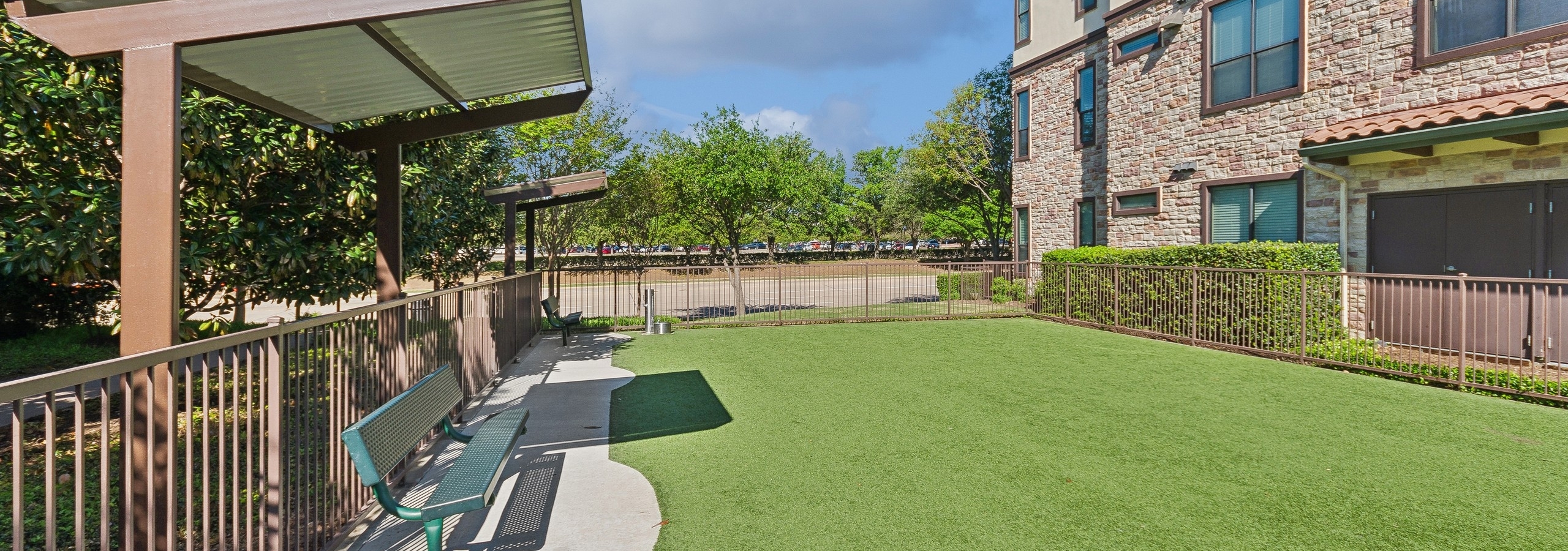 AMLI at Escena dog park with green turf and shaded green benches surrounded by a brown metal fence with trees and a stone apartment building