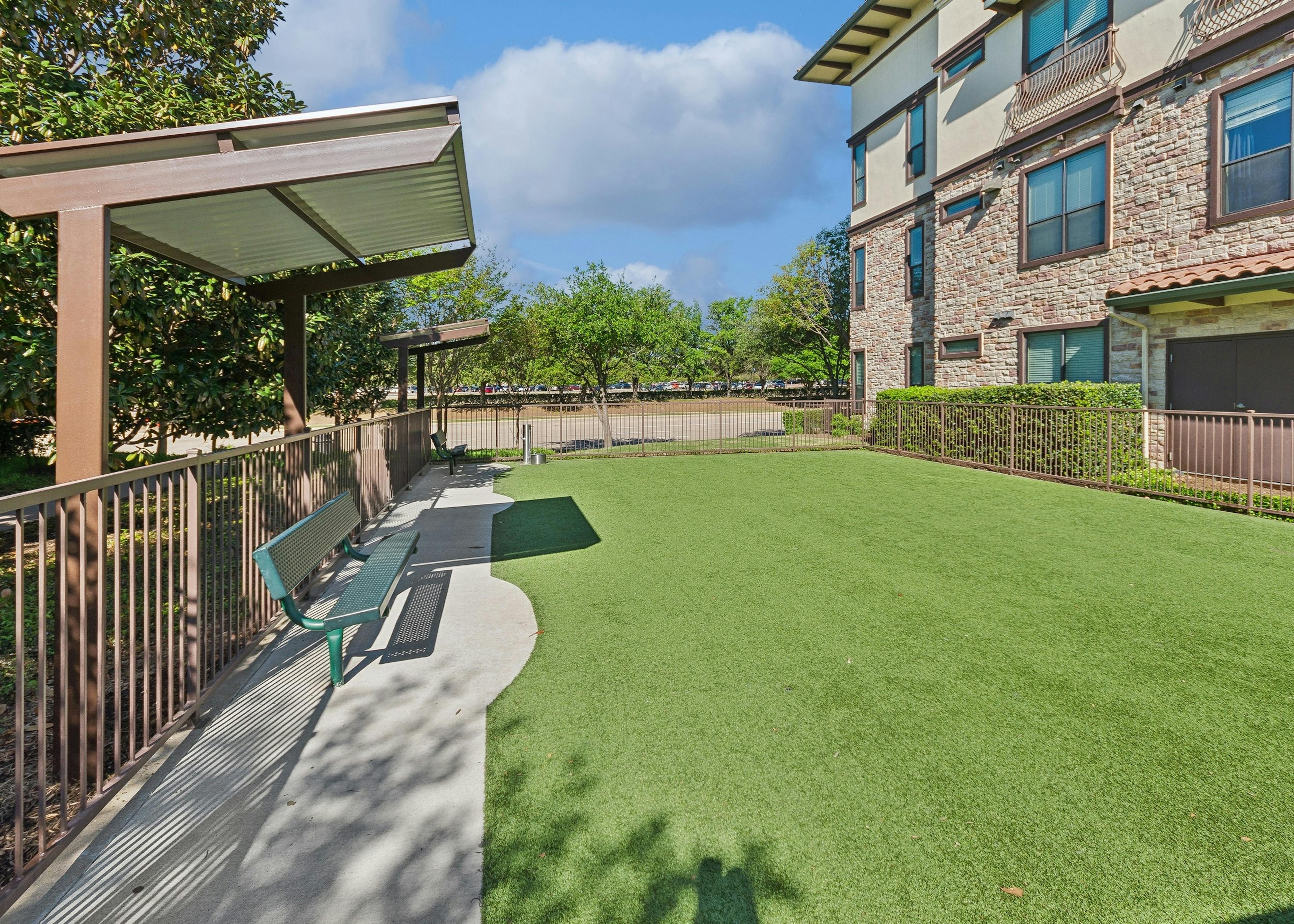 AMLI at Escena dog park with green turf and shaded green benches surrounded by a brown metal fence with trees and a stone apartment building