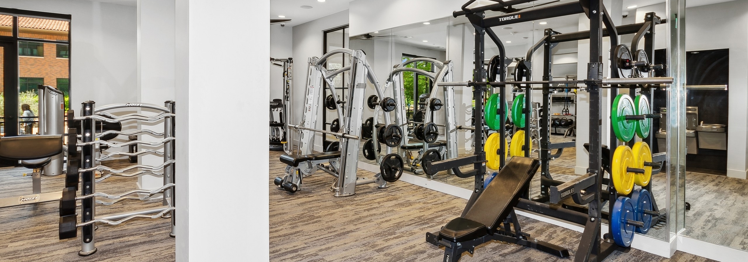 Carpeted gym at AMLI Addison with a mirrored wall with strength equipment including two large weight benches and weights
