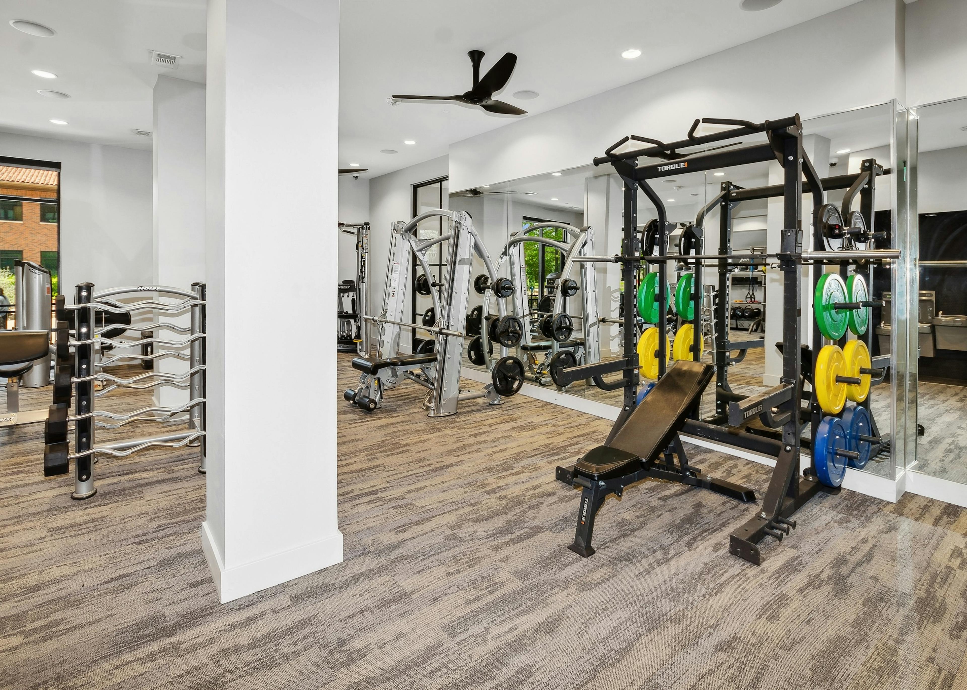 Carpeted gym at AMLI Addison with a mirrored wall with strength equipment including two large weight benches and weights 