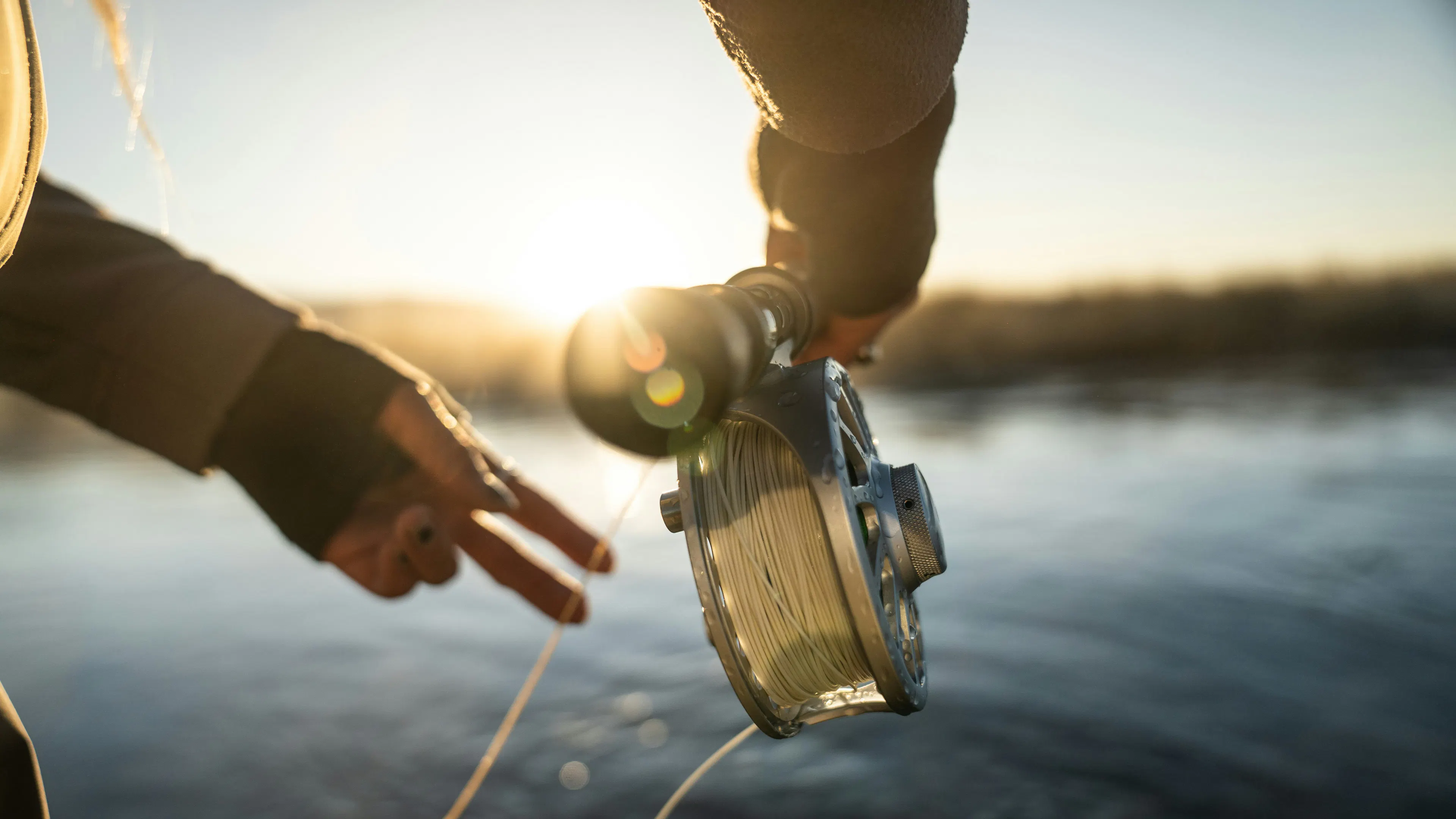 A close up of someone's hands holding and casting a fly fishing pole over a wide river