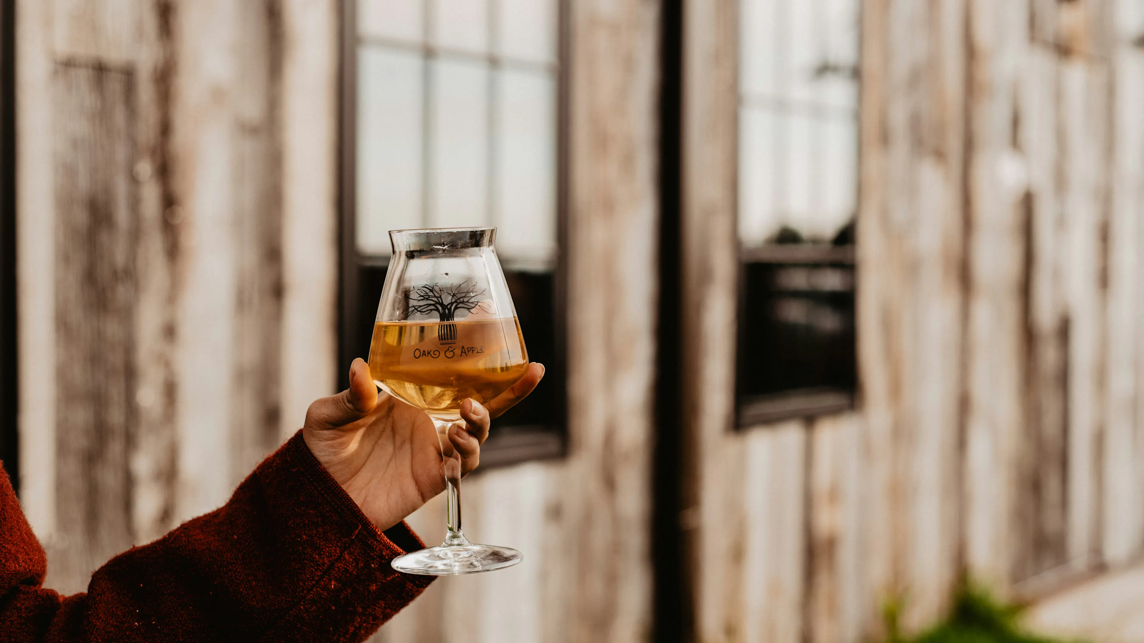 A woman's hand holding a glass of hard apple cider with a barn in the background