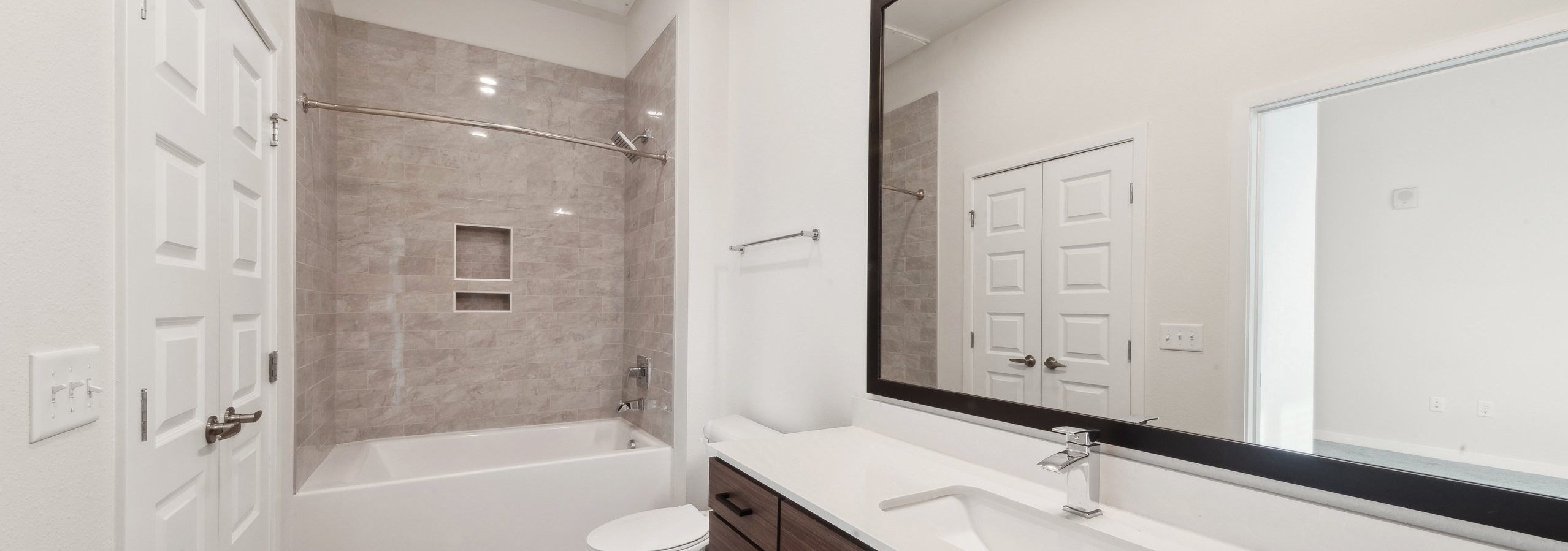 Bathroom at AMLI Art District featuring a large mirror over a brown vanity with white countertop and a white bathtub with beige tile walls
