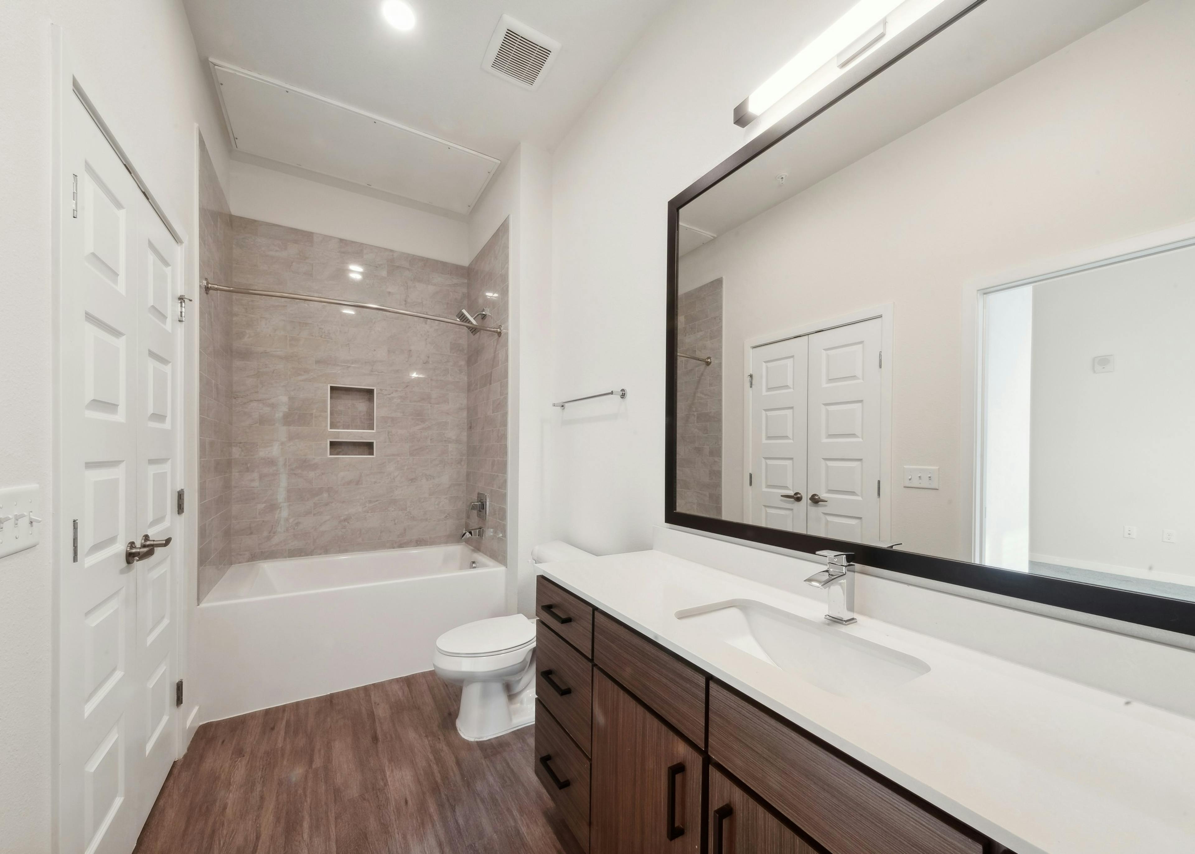 Bathroom at AMLI Art District featuring a large mirror over a brown vanity with white countertop and a white bathtub with beige tile walls
