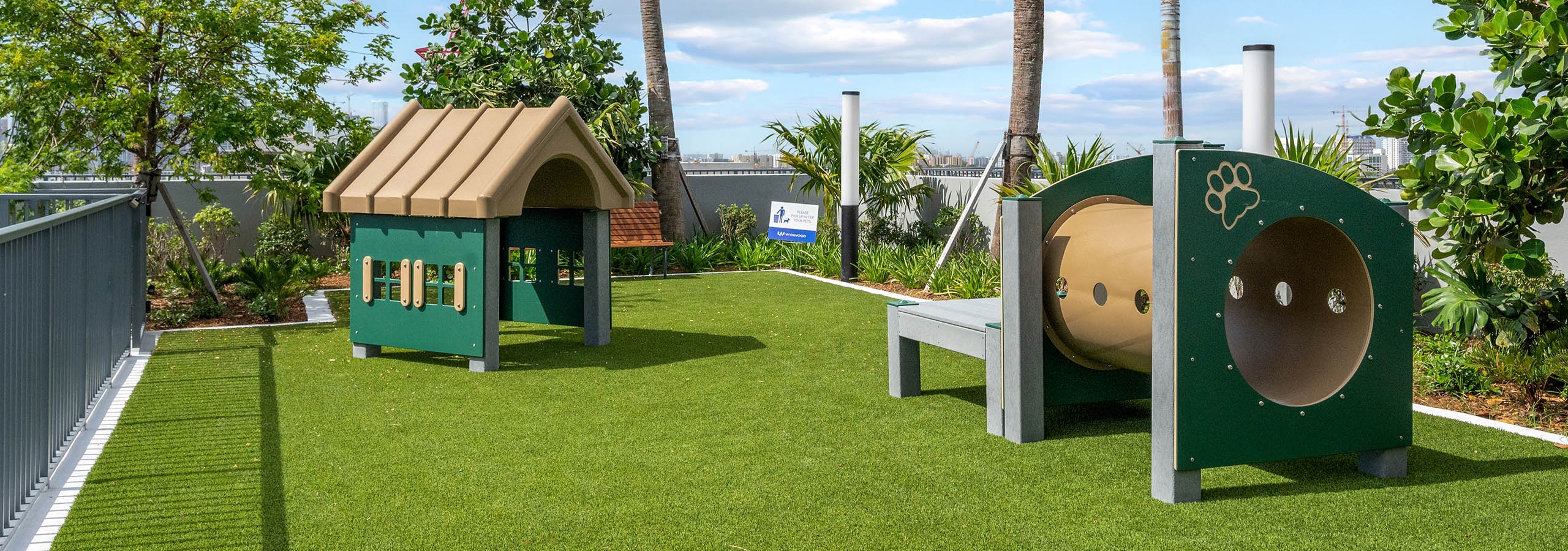 A daytime view of the rooftop gated pet park at AMLI Wynwood apartments surrounded by trees with green turf and two play tunnels